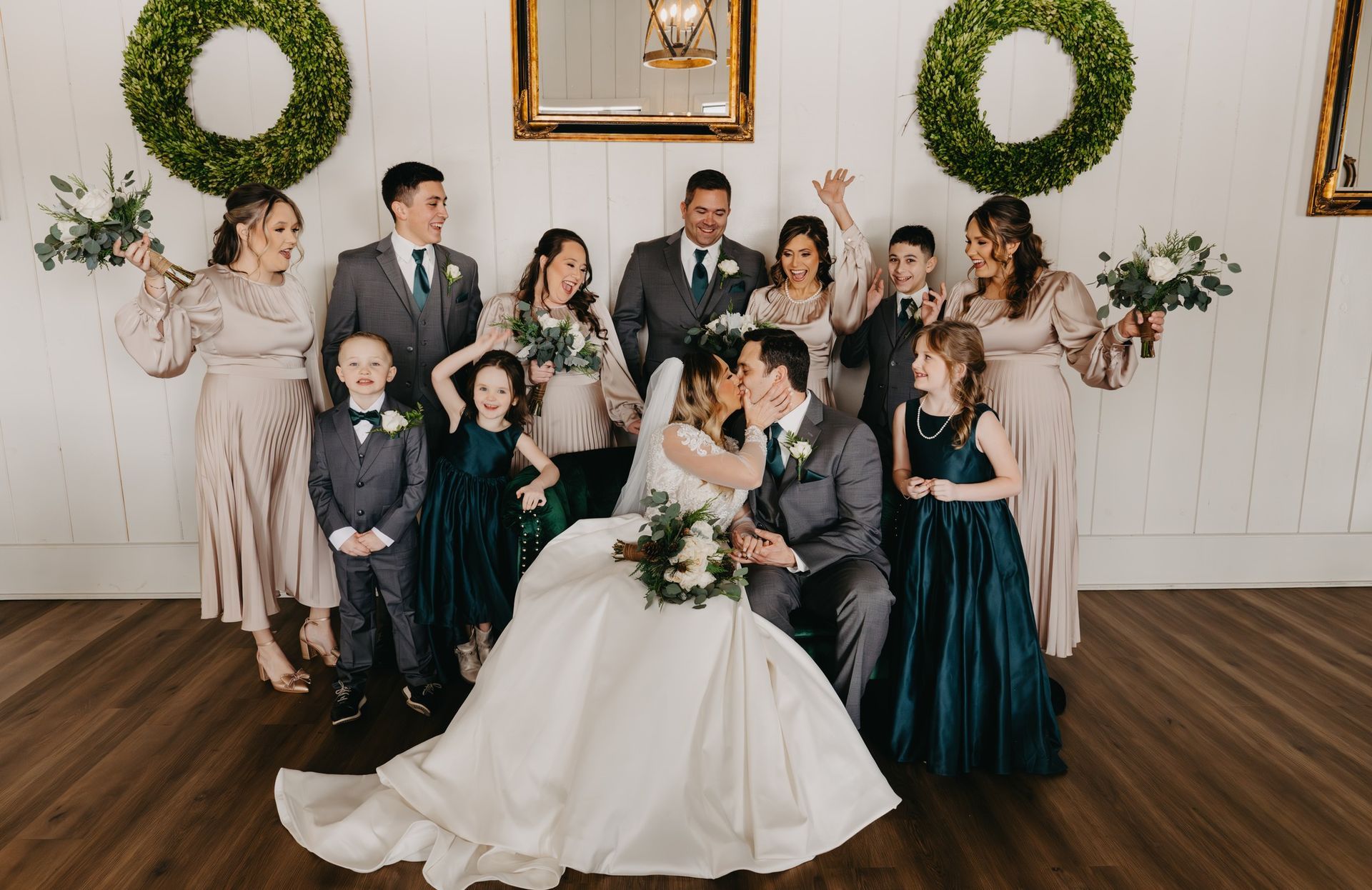 A wedding party posing for a group portrait indoors, featuring the couple kissing in front of friends and family.