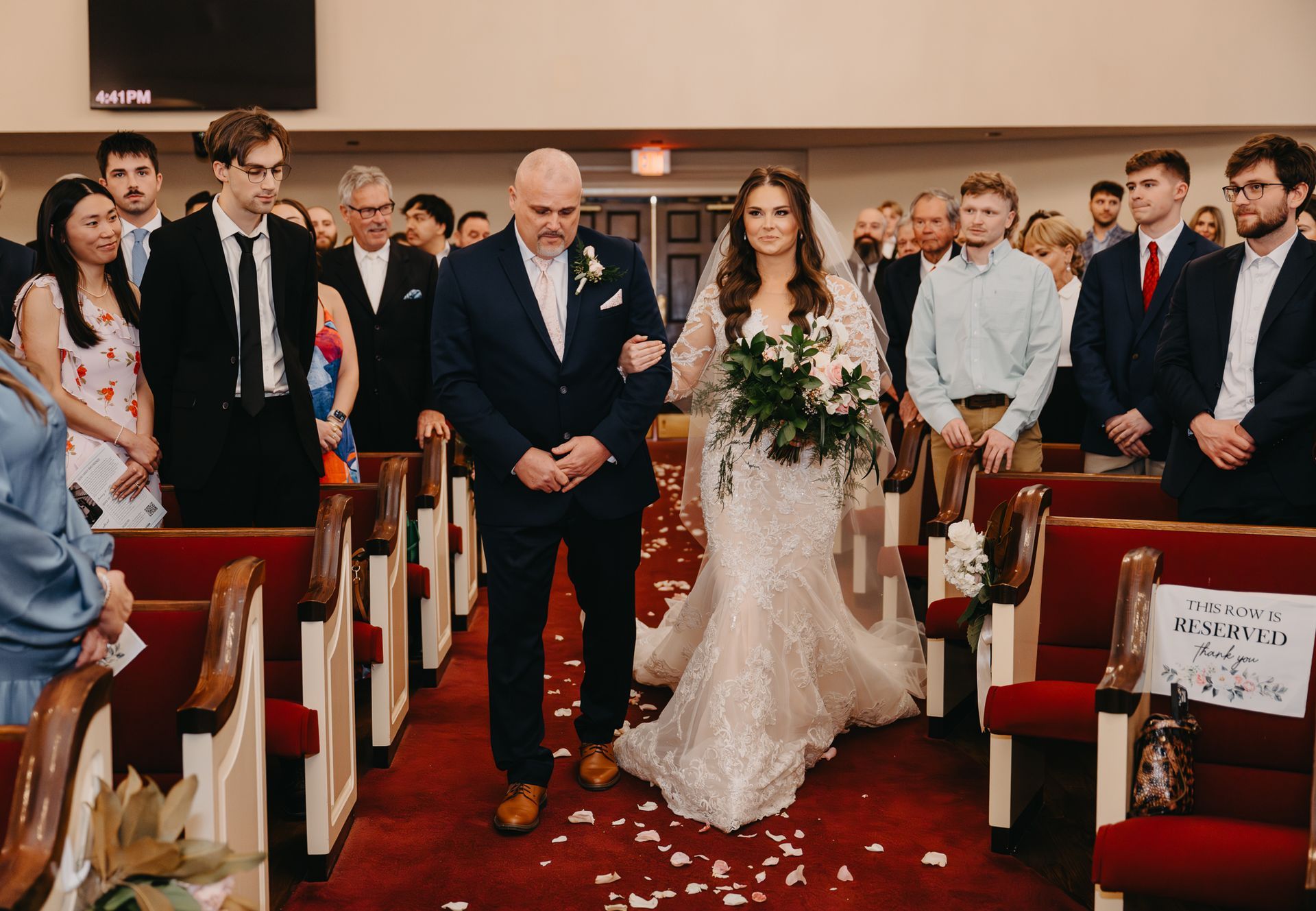 A wedding procession: a person in a suit and a person in a white gown walking down a red aisle among guests in a church.