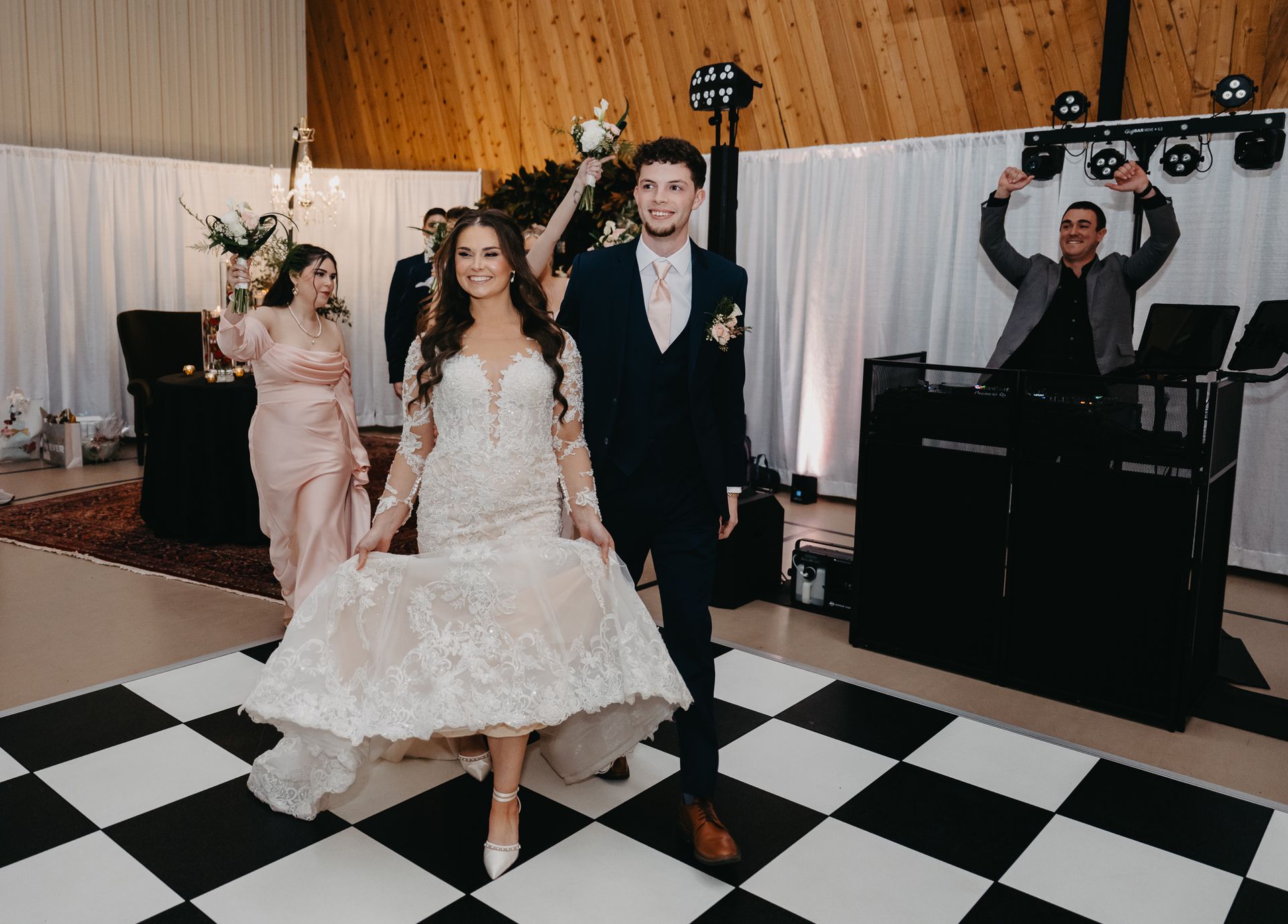 A bride in a lace gown and a groom in a navy suit walk together on a black-and-white checkered dance floor at a wedding.
