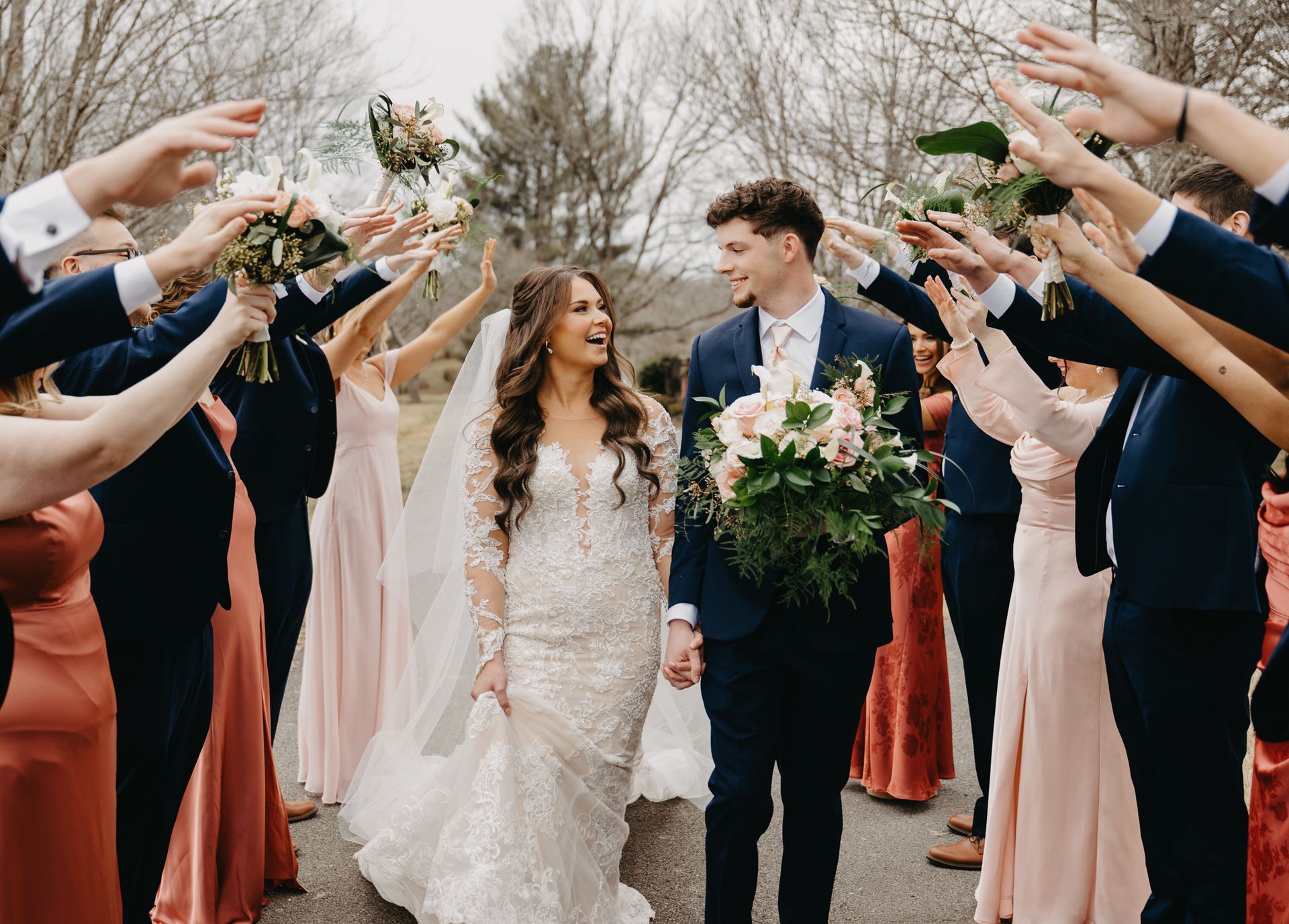 A smiling bride and groom walk through a tunnel of hands held by a wedding party in a park setting.