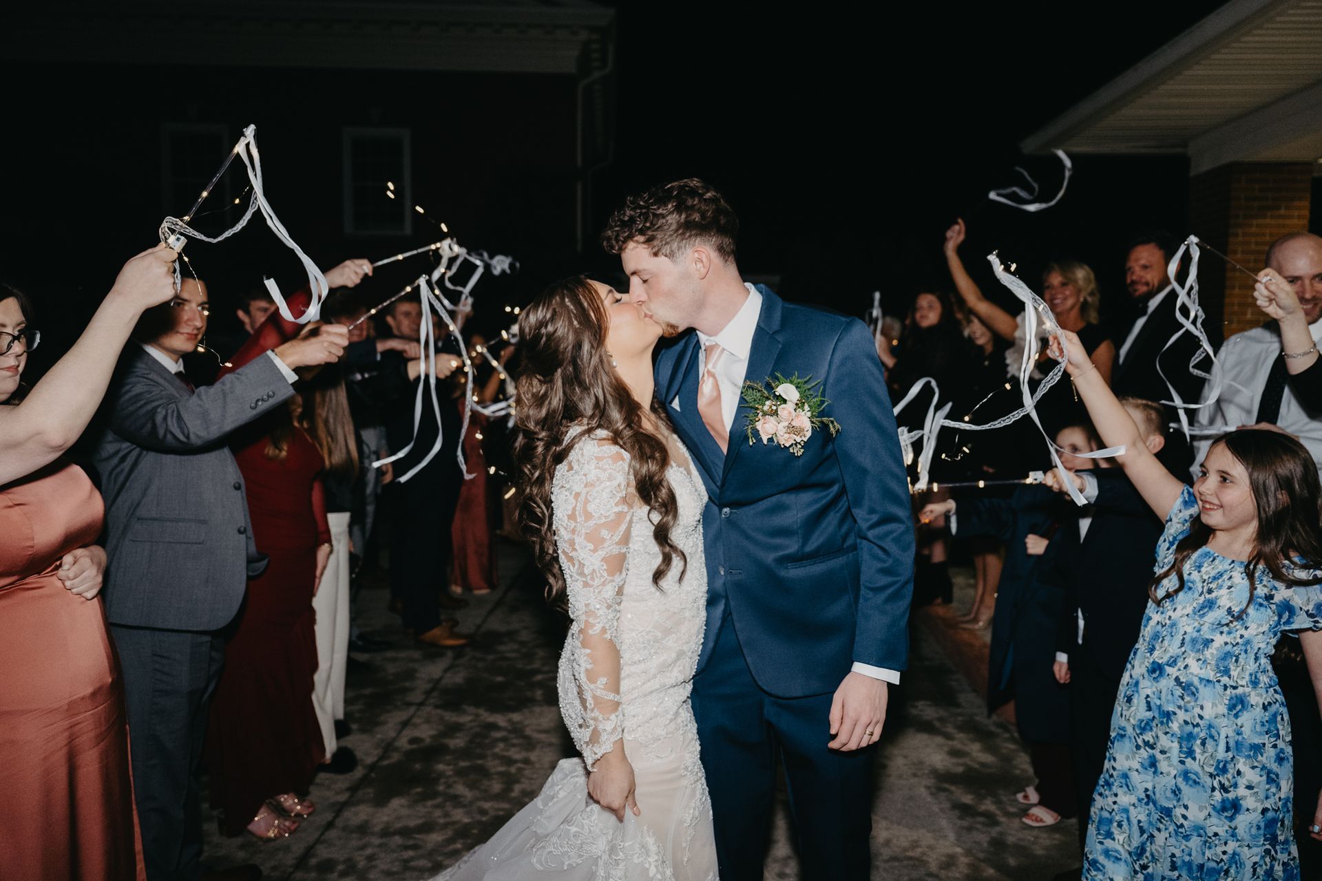A newly married couple kisses while guests wave white ribbon wands at an outdoor evening wedding exit.