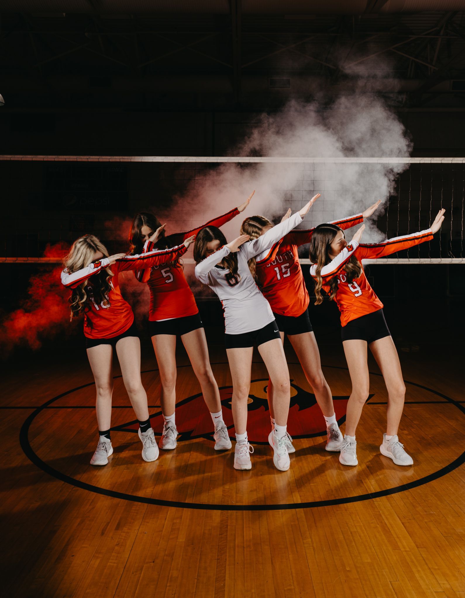 Five volleyball players in uniforms stand on a court, performing a synchronized dab with smoke effects in the background.