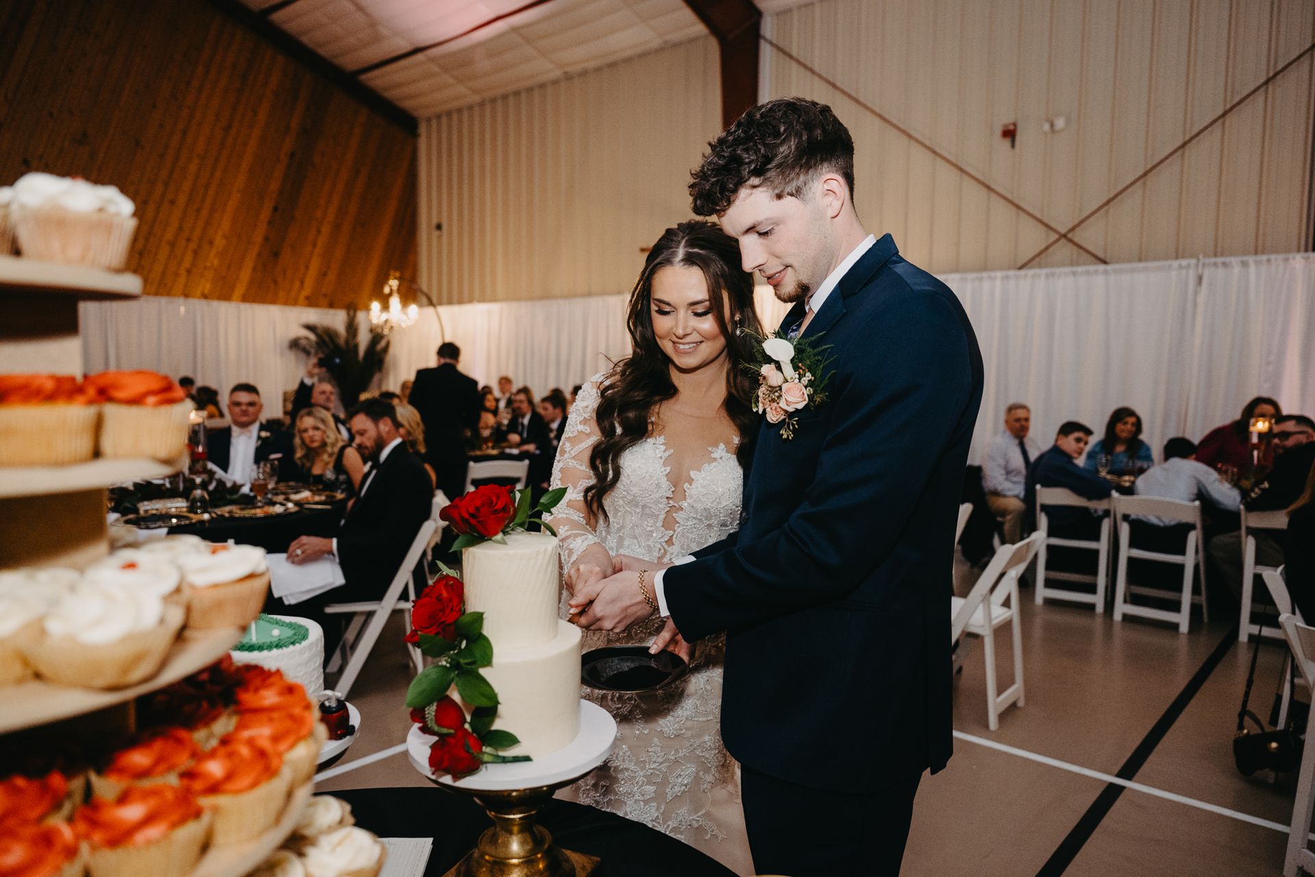 A couple in wedding attire cuts a tiered cake decorated with red roses at an indoor reception.