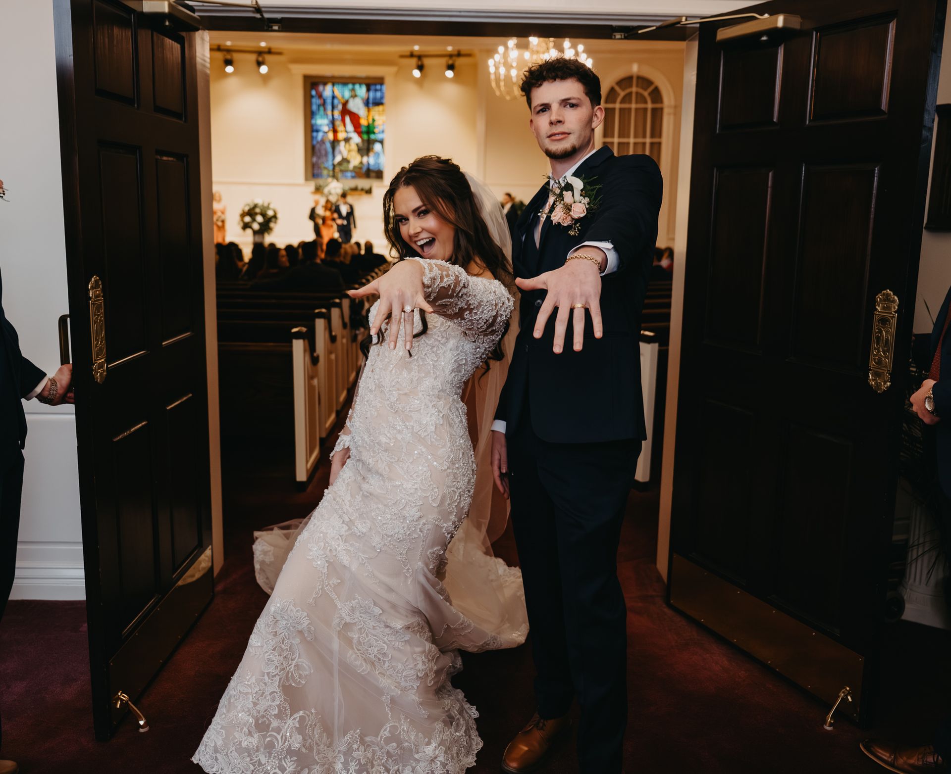 A bride and groom posing in a church doorway, smiling and excitedly displaying their wedding rings to the camera.