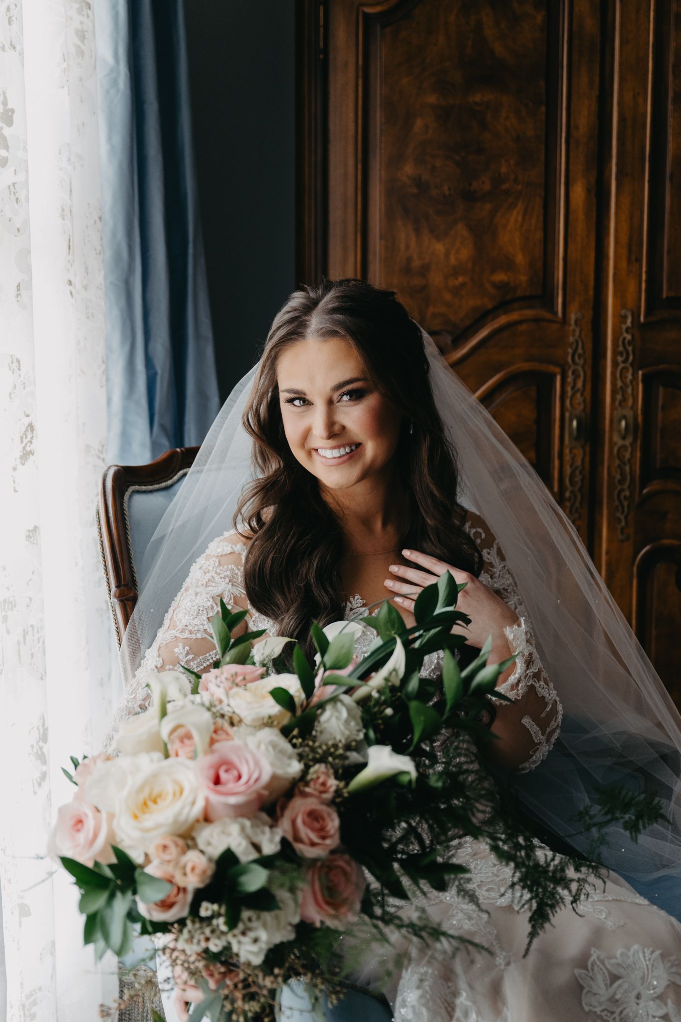 A bride in a lace gown and veil smiles while holding a bouquet of pink and white flowers, seated in a wooden chair.
