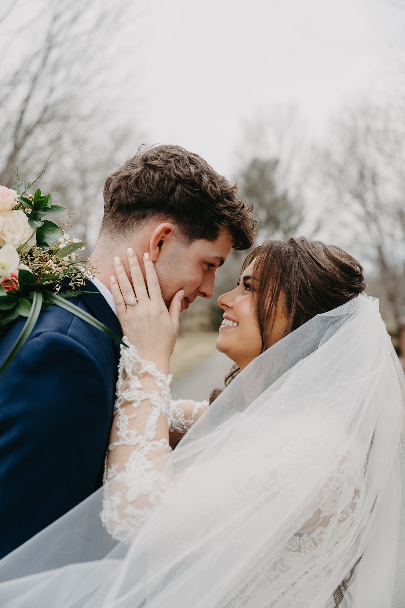 A bride in a lace gown and veil gazes lovingly at her groom, who holds her, outdoors against a backdrop of bare trees.