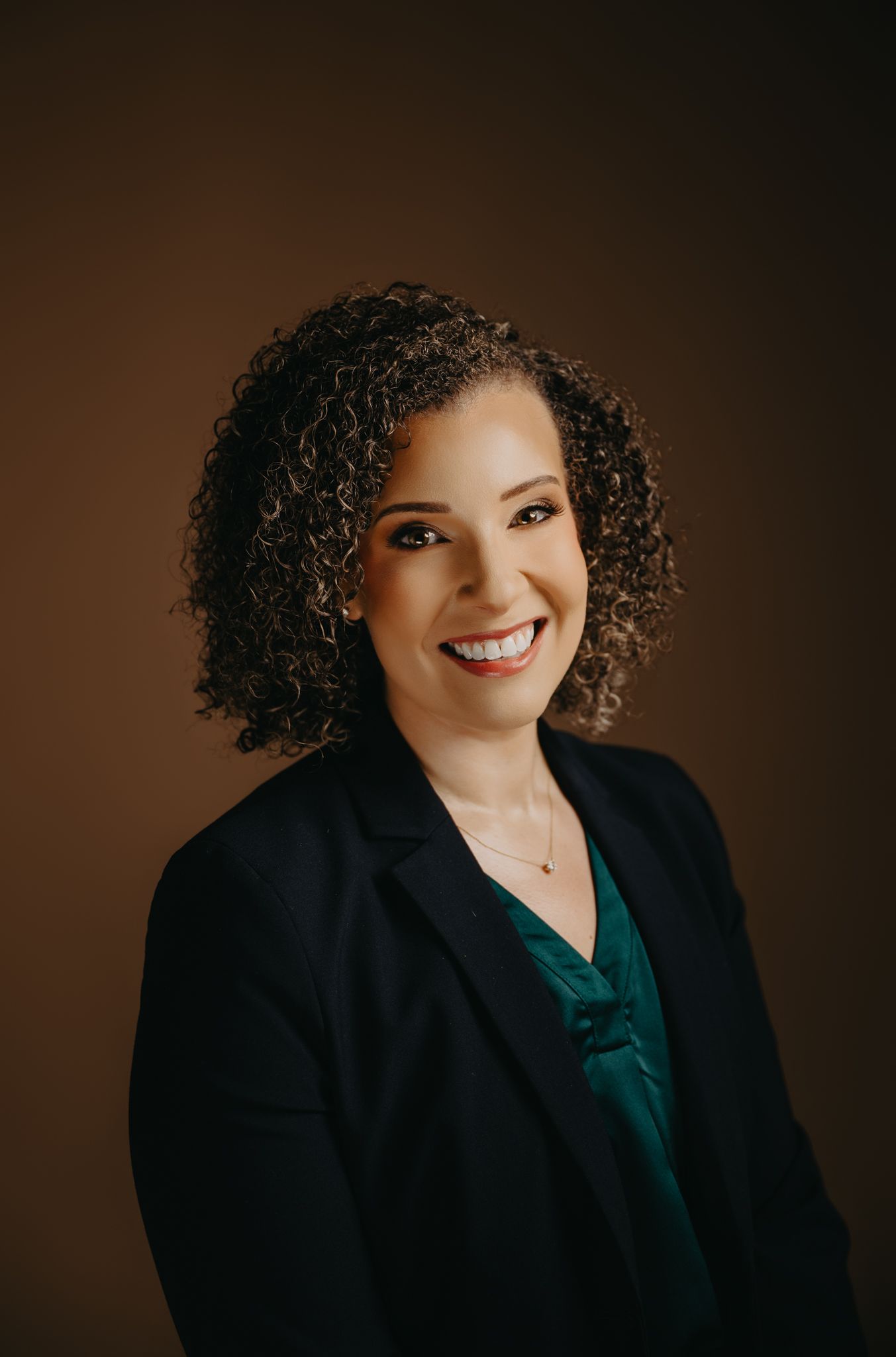 Professional headshot of a smiling person with curly dark hair, wearing a green blouse and dark blazer.
