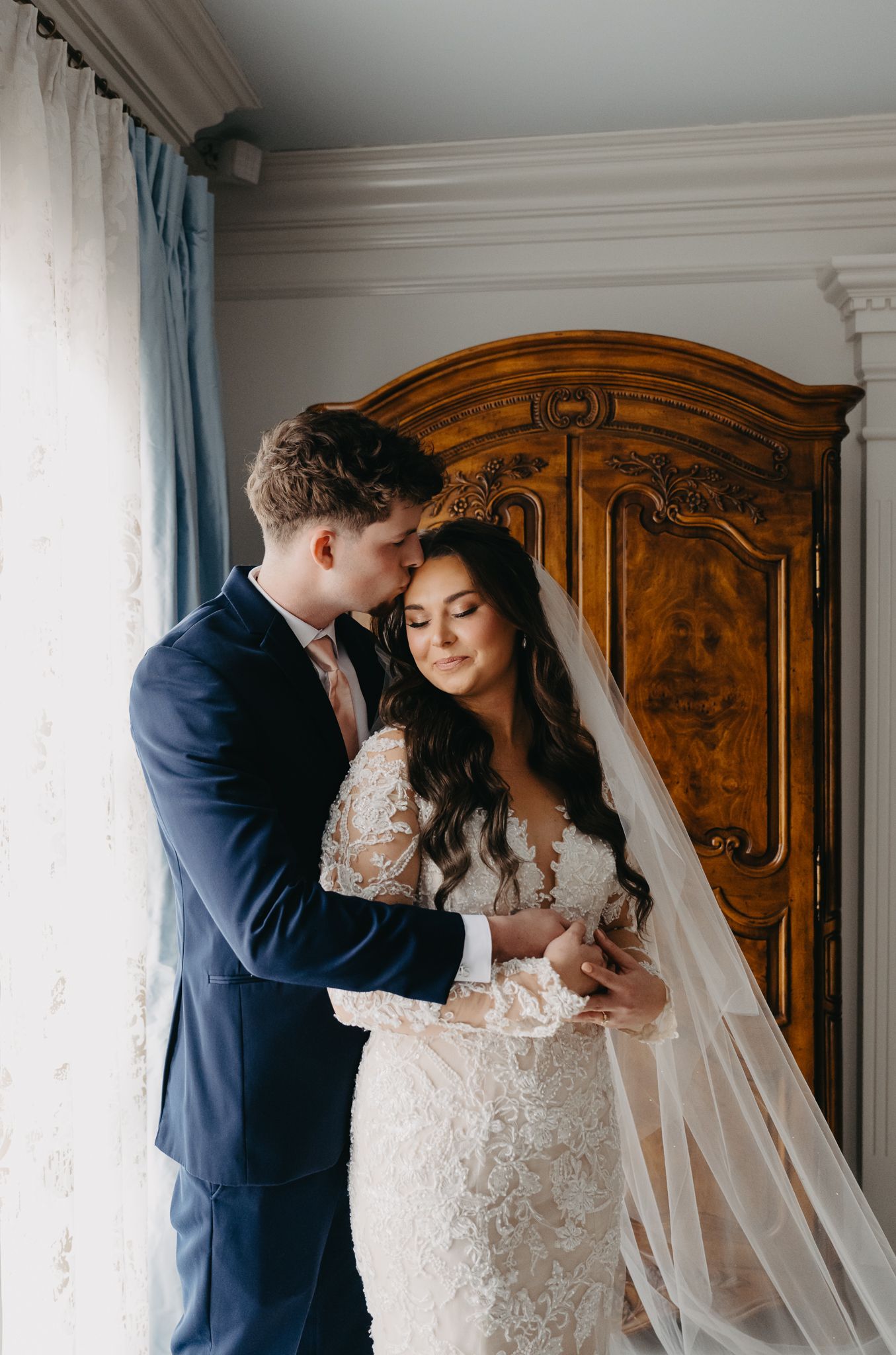 A groom in a dark suit kisses the bride on her forehead as she wears a long, lacy white gown with a flowing veil.