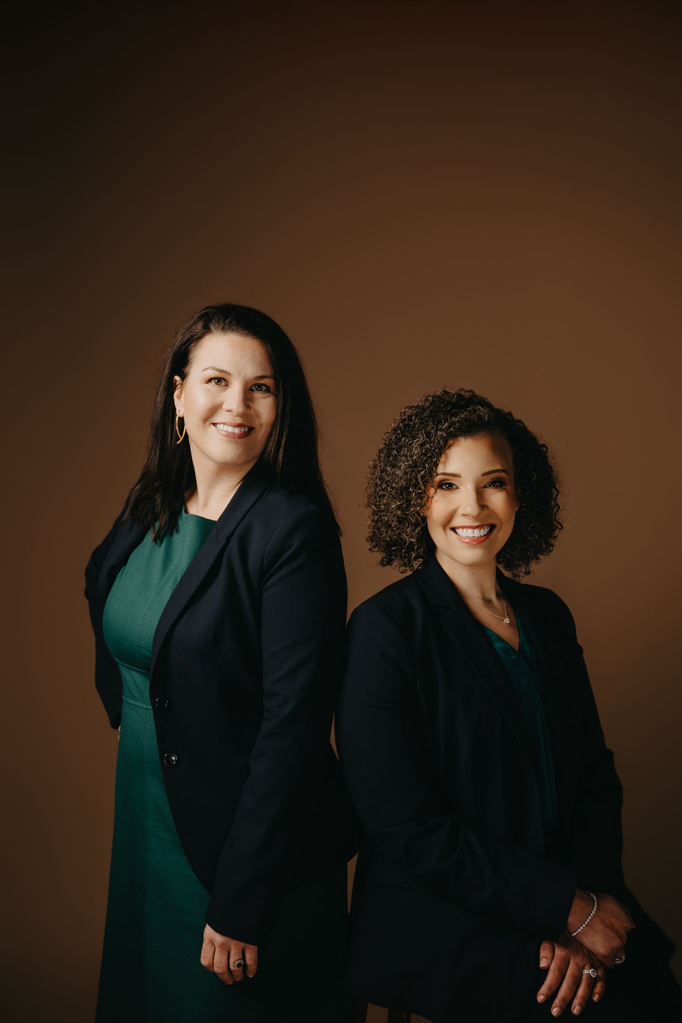 Two smiling people stand against a brown background, both wearing dark blazers over green tops.