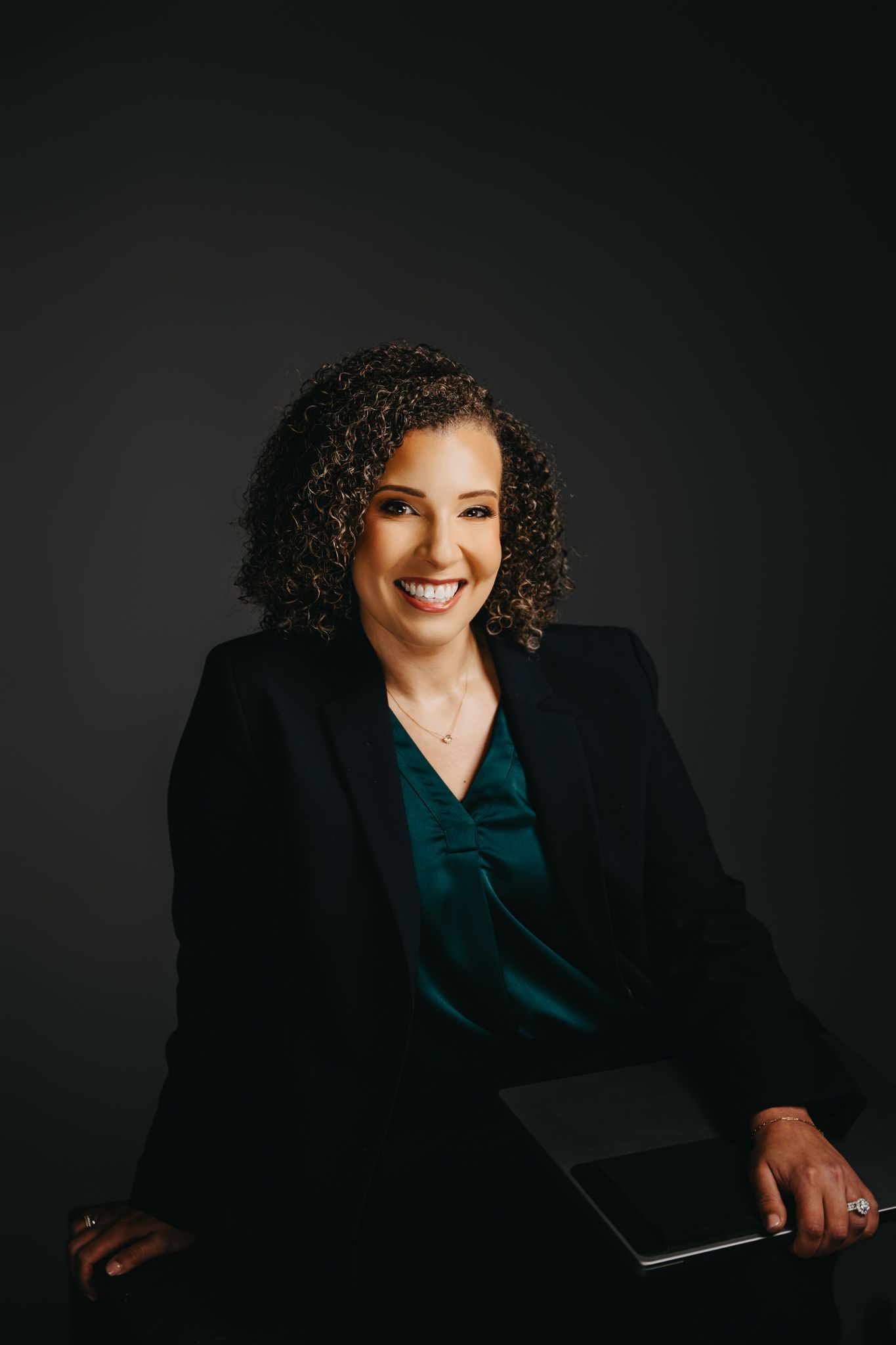 A smiling professional in a dark blazer and green blouse sits against a dark background, holding a small black portfolio.