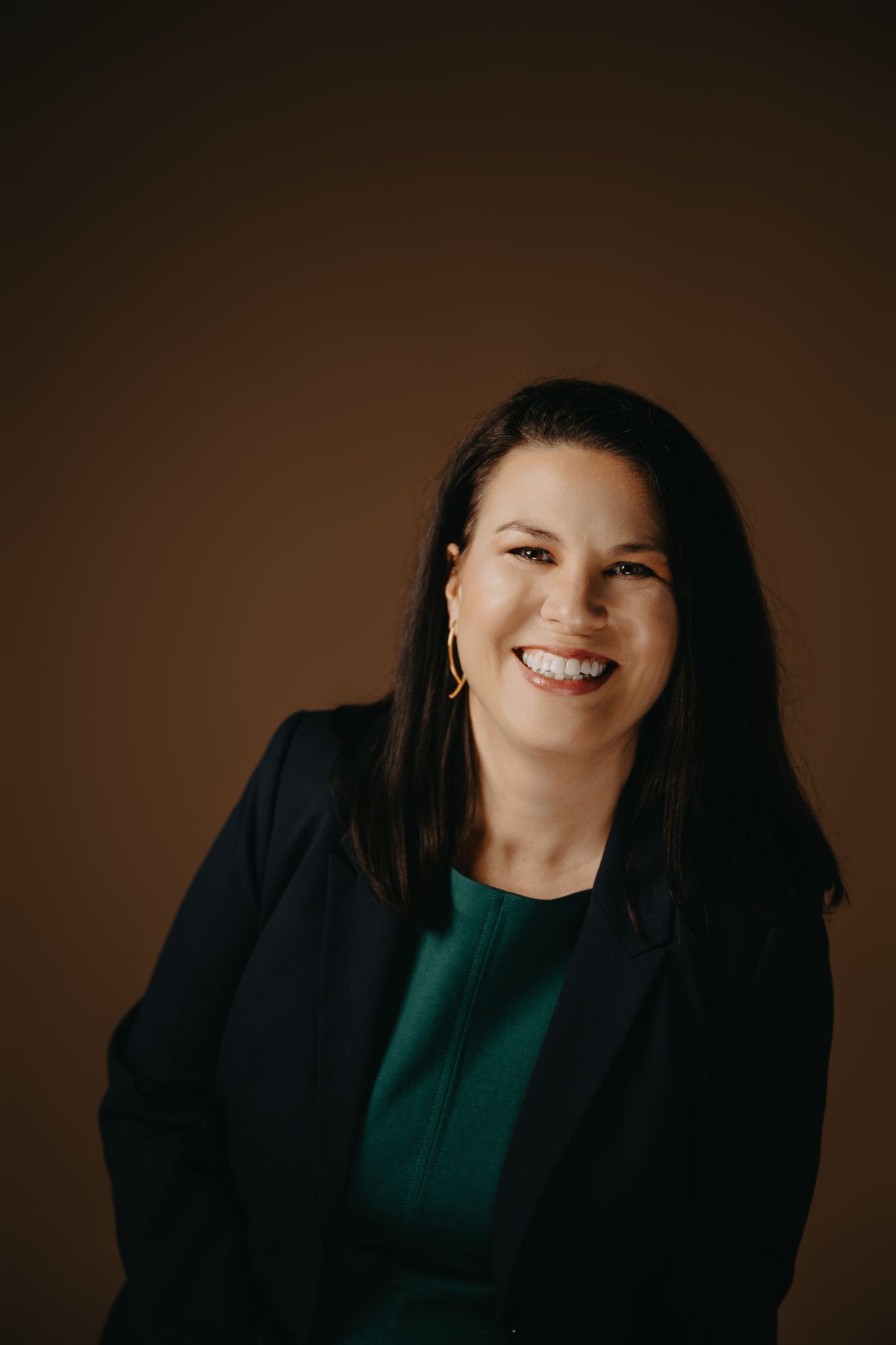 Smiling person with dark hair wearing a dark blazer over a green top against a solid brown background.