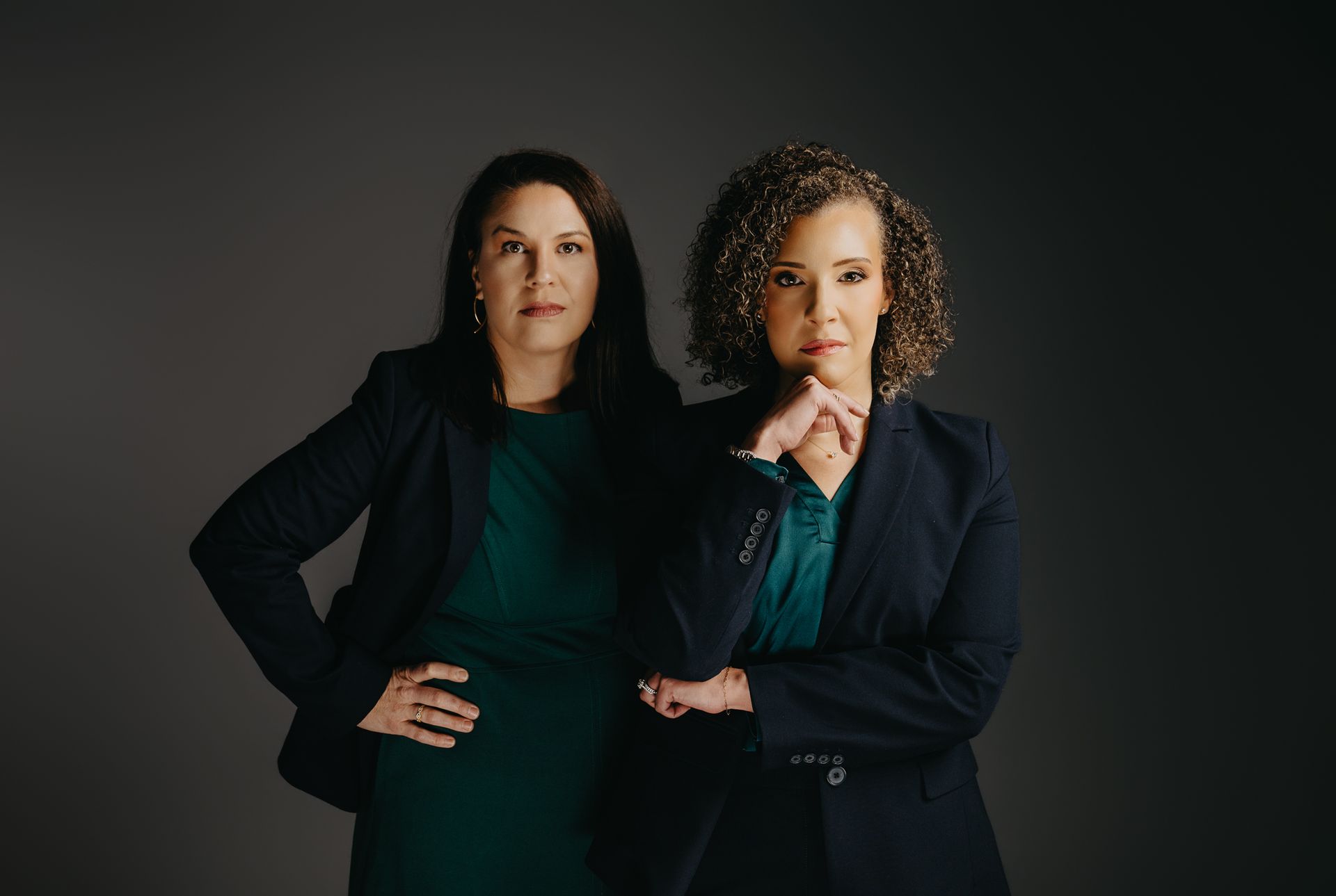 Two professionals in dark blazers and green tops pose against a gray background with neutral expressions.
