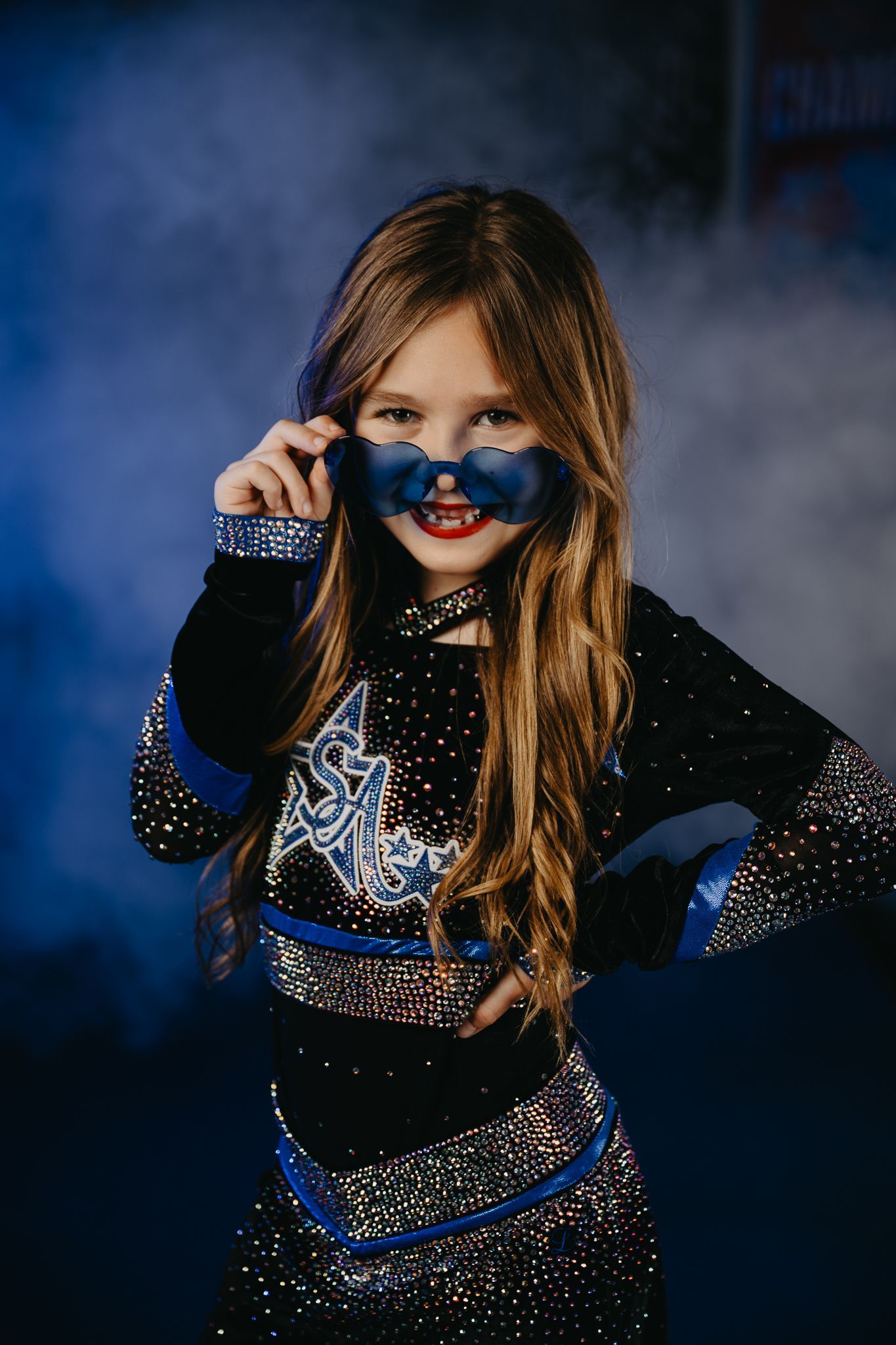 A young person in a glittery blue and black outfit looks through blue heart-shaped glasses amidst blue swirling smoke.