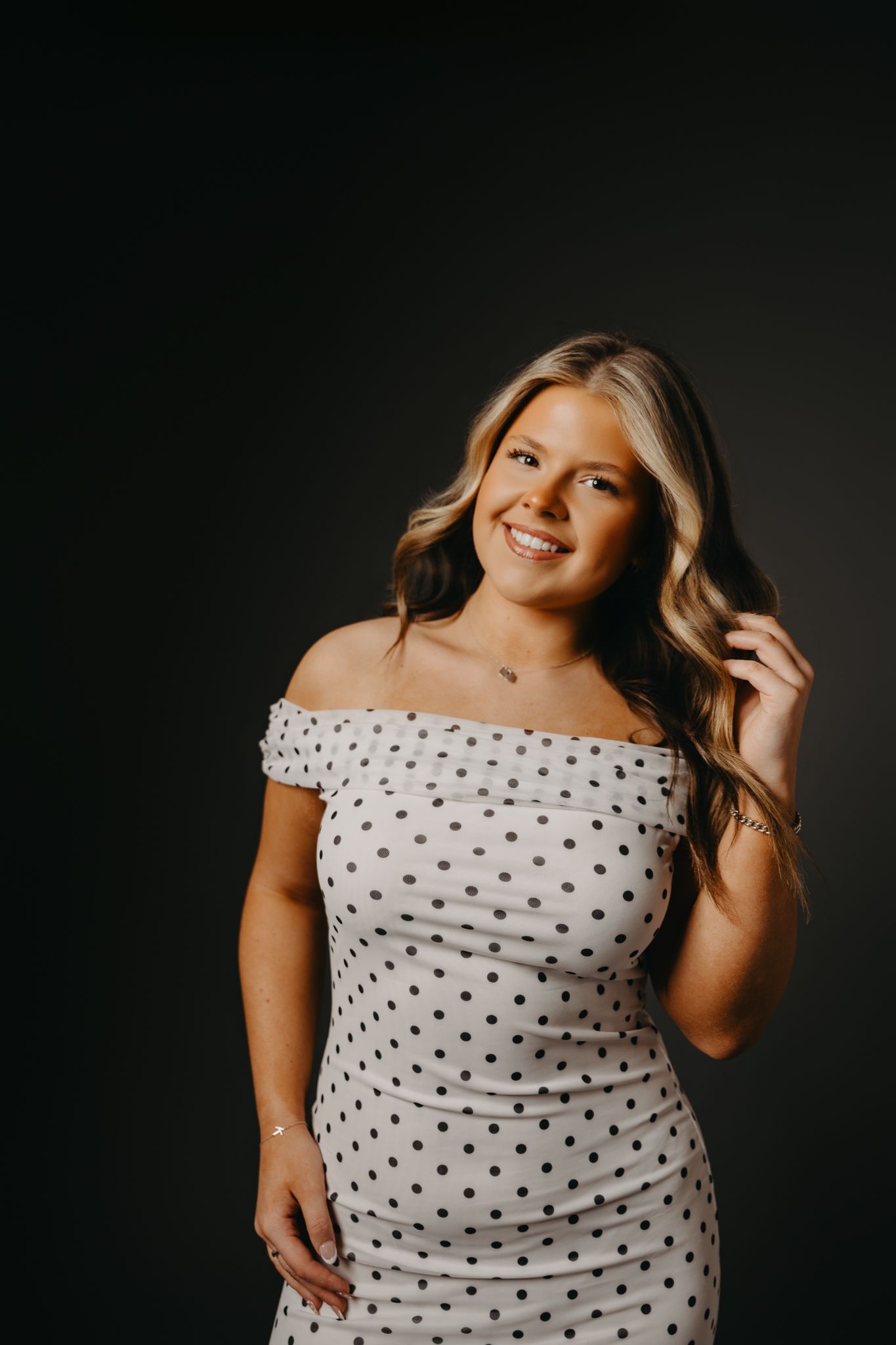 A smiling person with wavy hair wears a white, off-the-shoulder polka-dot dress against a dark background.