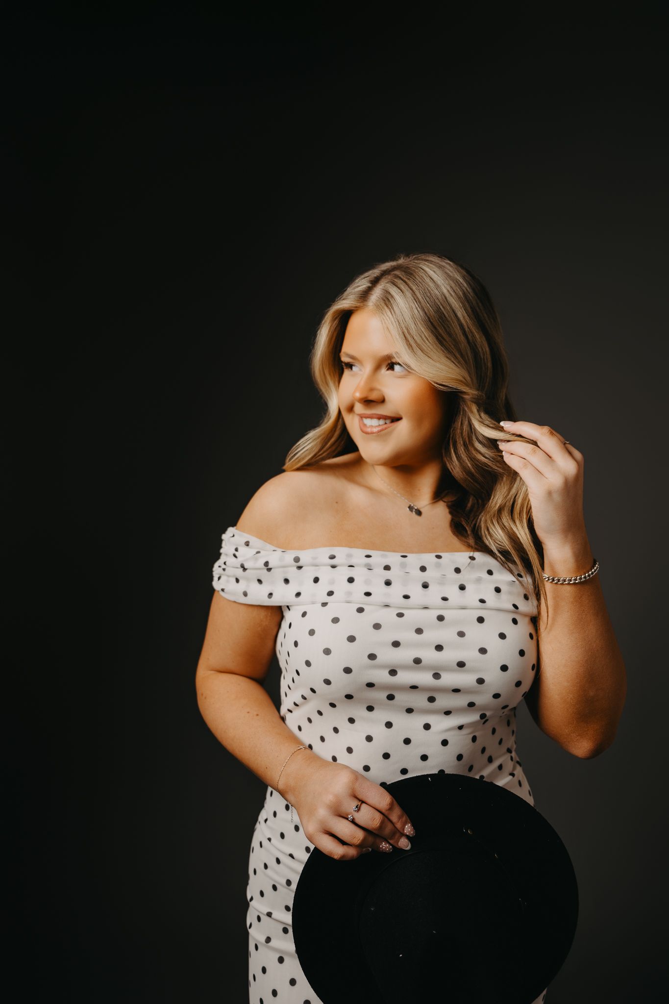 A person with blonde hair smiles while holding a black hat, wearing a white off-the-shoulder dress with black dots.