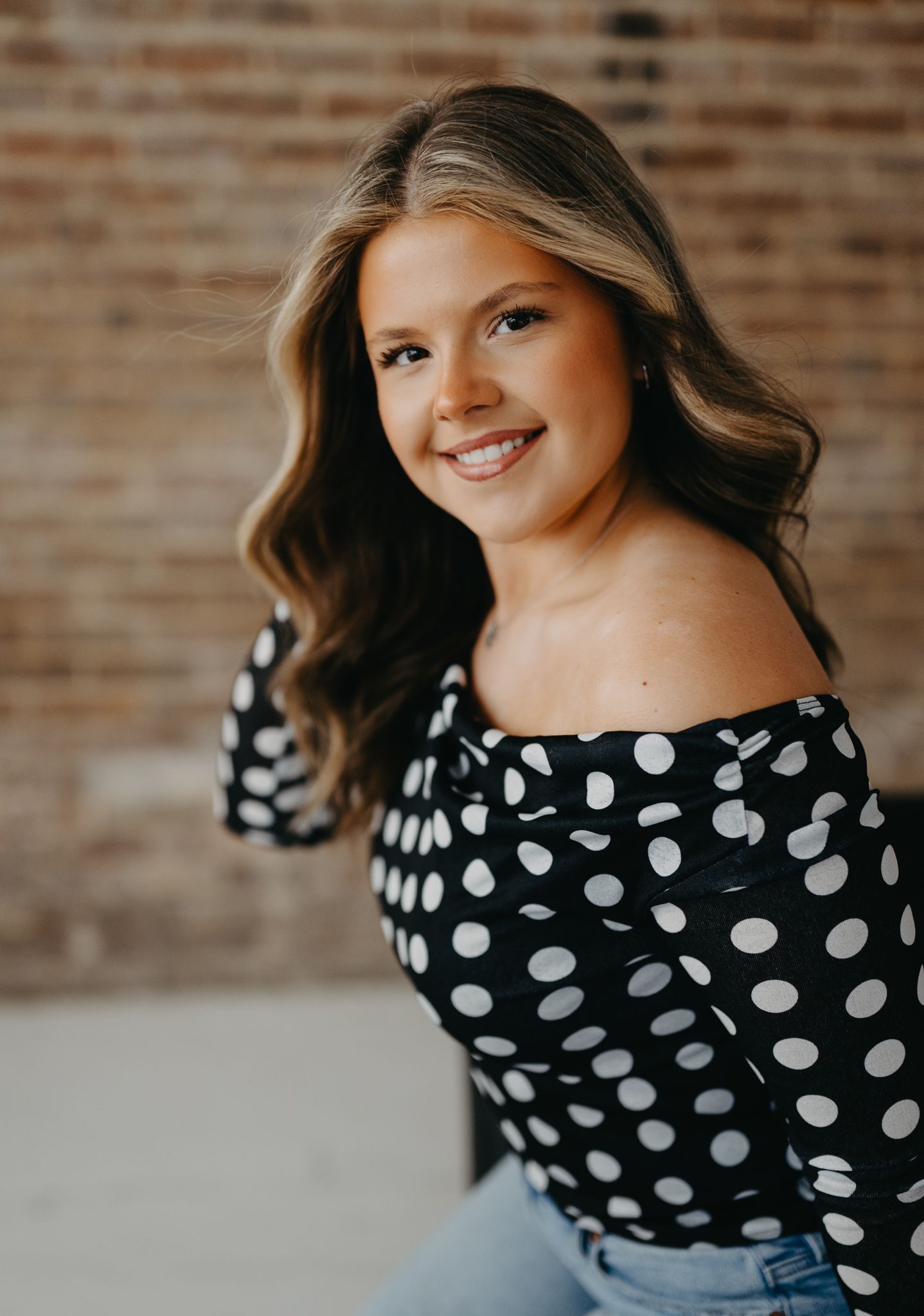 A smiling person wearing a black and white polka dot off-the-shoulder top poses against a textured brick wall background.