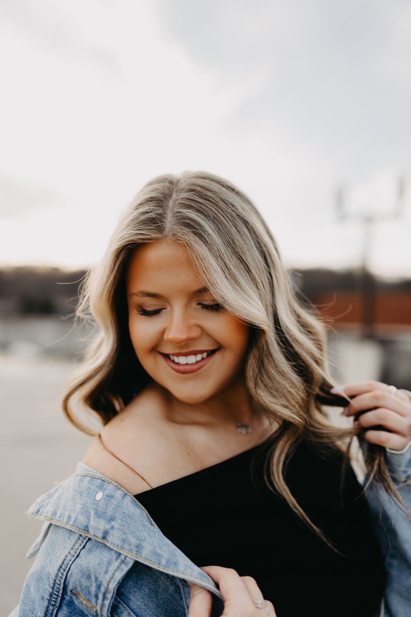 A person smiling with wavy blonde hair, wearing a black top and denim jacket, looking downward against a blurred background.
