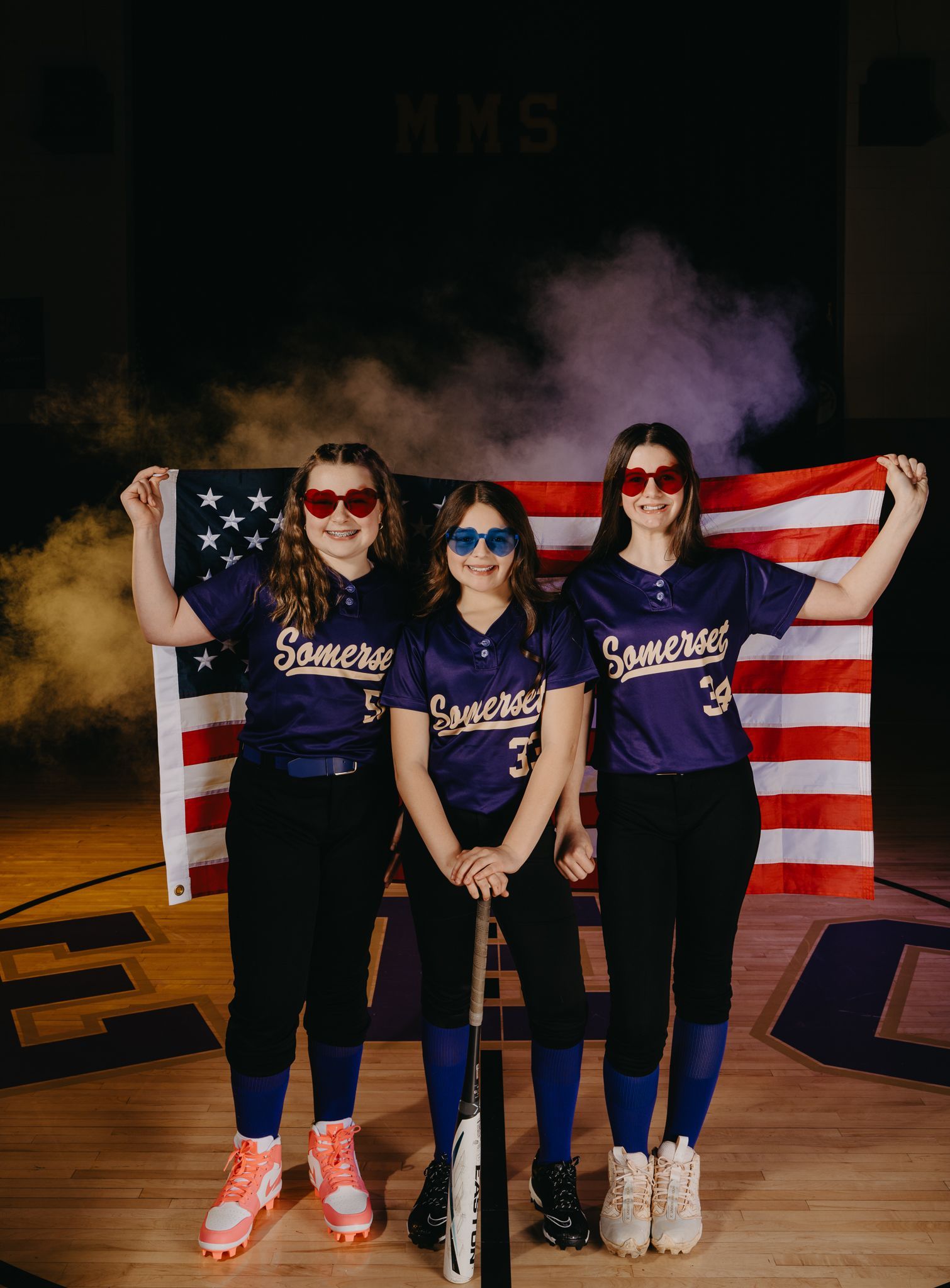 Three athletes in matching purple jerseys and sunglasses stand on a gym floor before an American flag amid purple smoke.