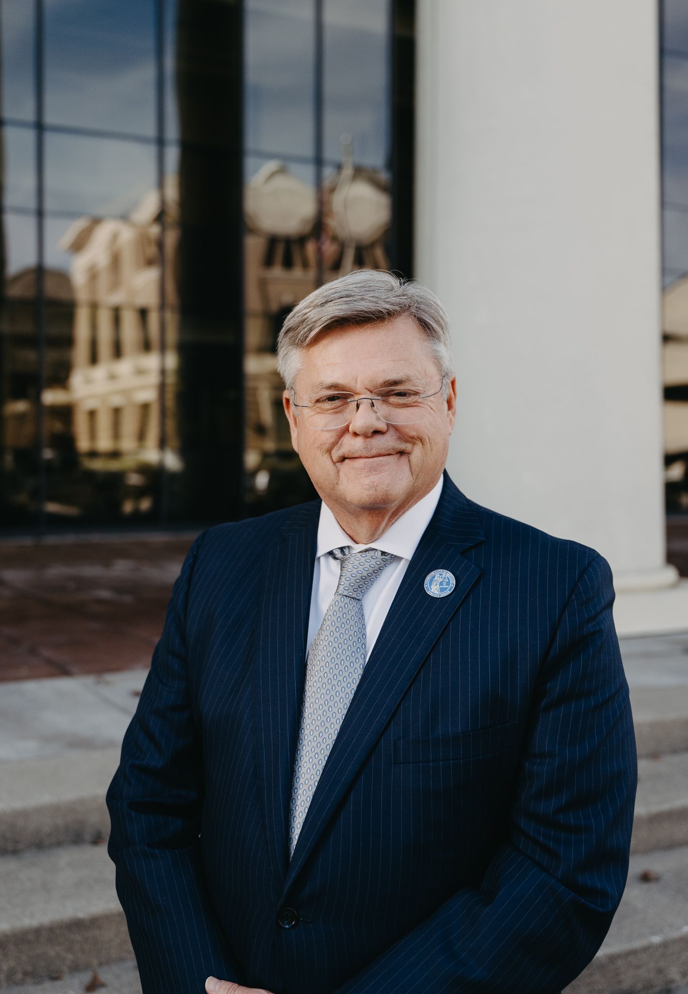 A man in a pinstriped navy suit and patterned tie poses for a portrait in front of a building with glass windows.