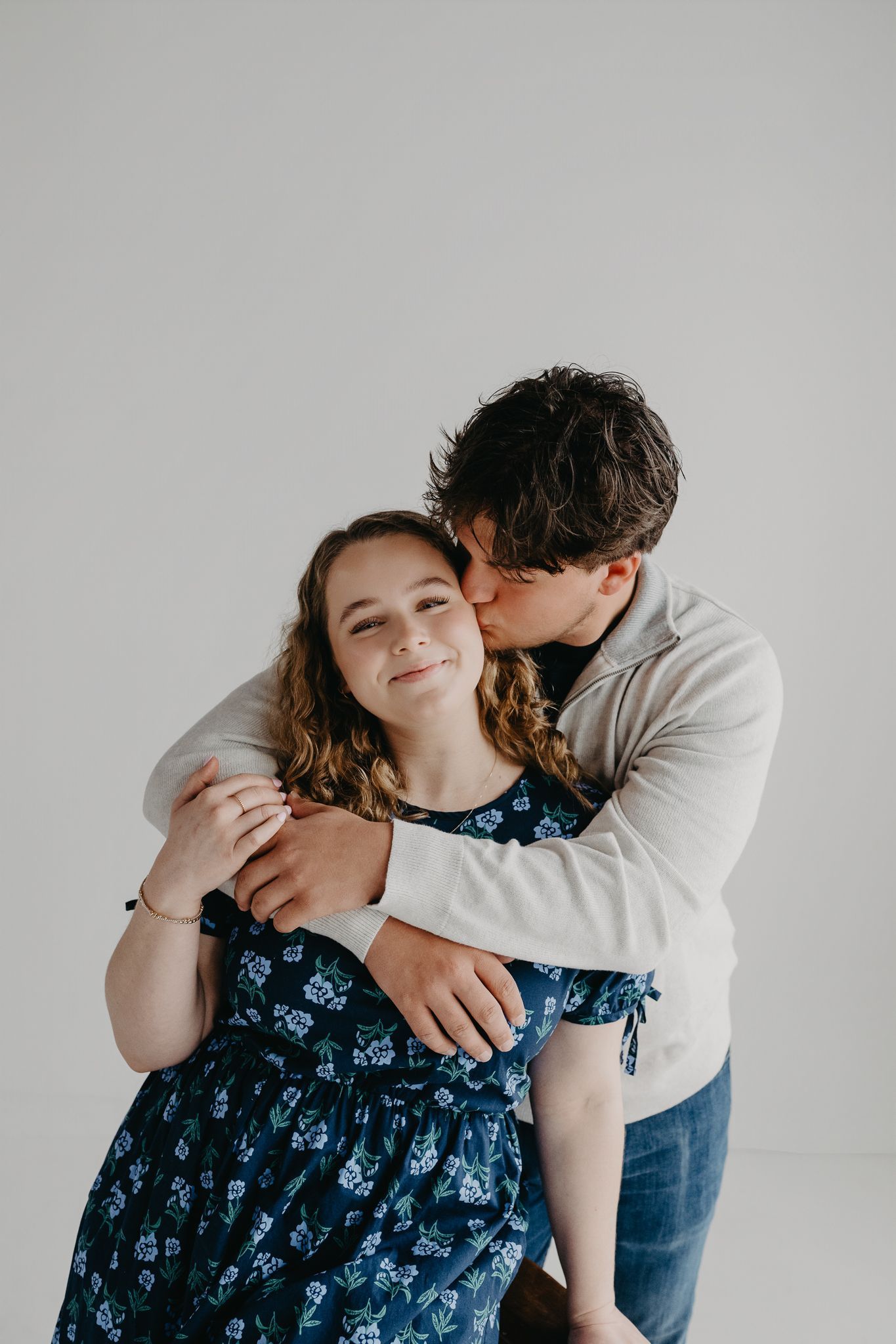 A person in a light sweater hugs someone in a blue floral dress from behind, kissing their cheek against a plain background.
