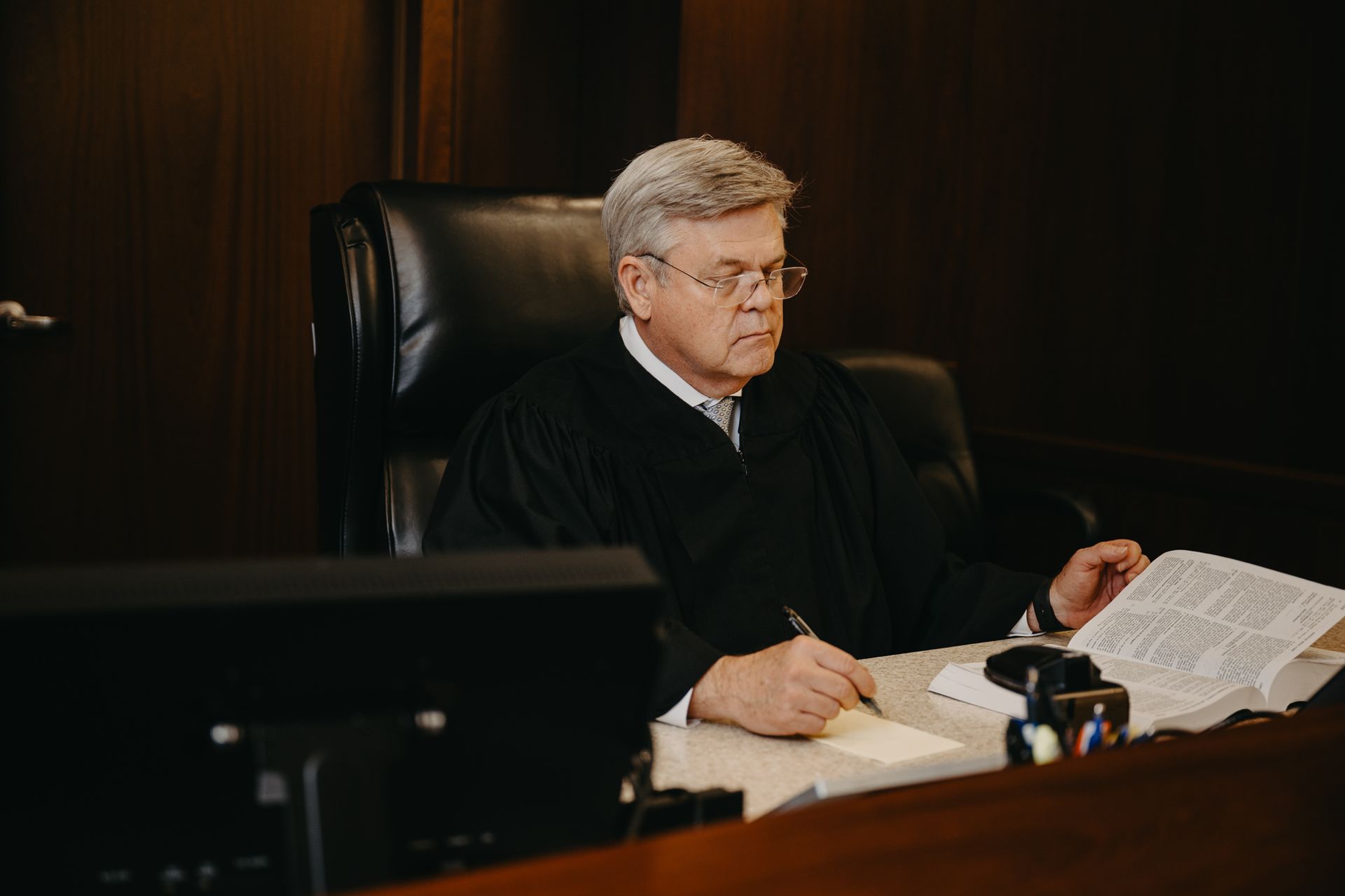 A judge in a black robe sits behind a desk in a courtroom, looking at a document and writing notes.