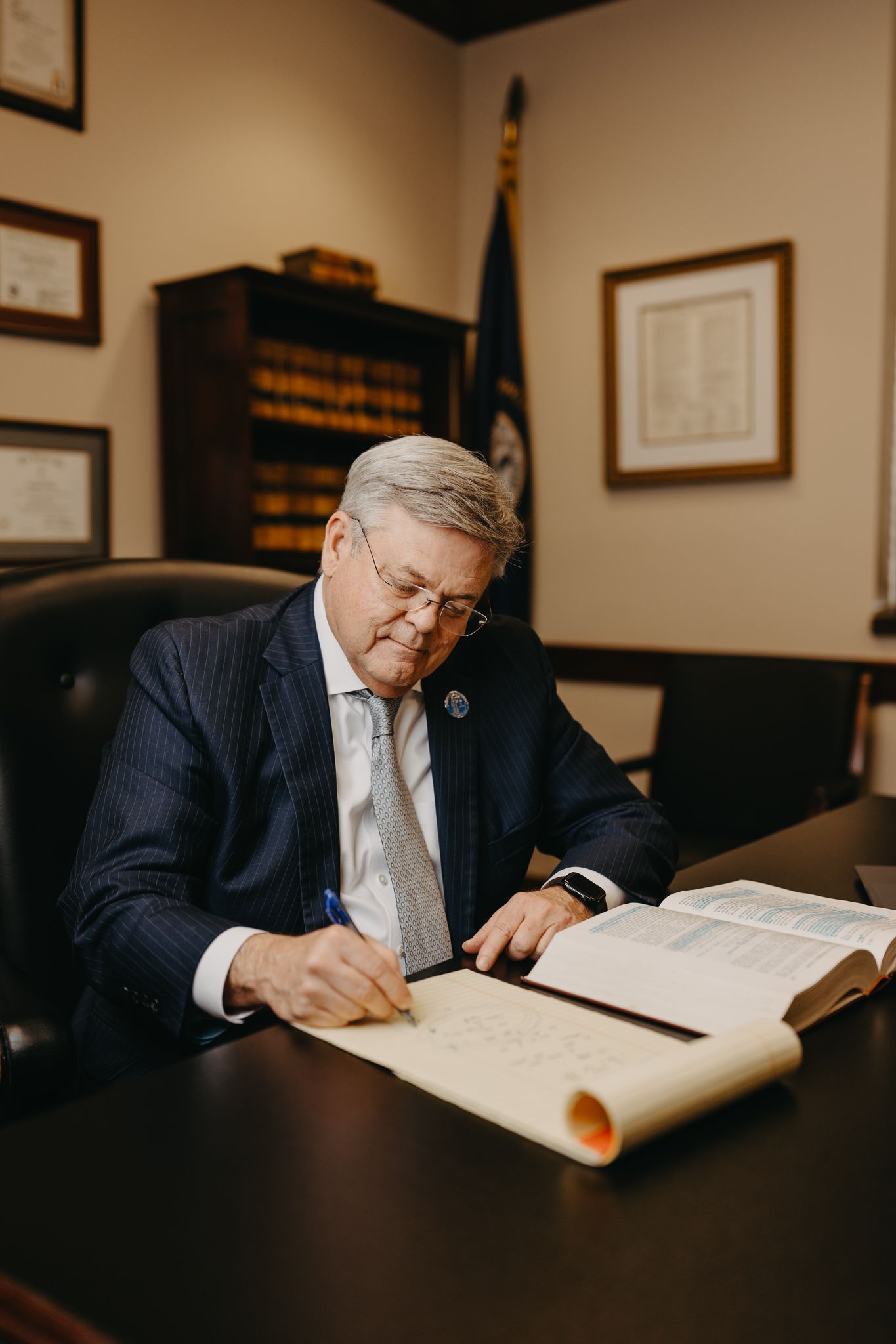 A professional sitting at a dark desk writing in a large book in an office with framed documents and a flag behind them.