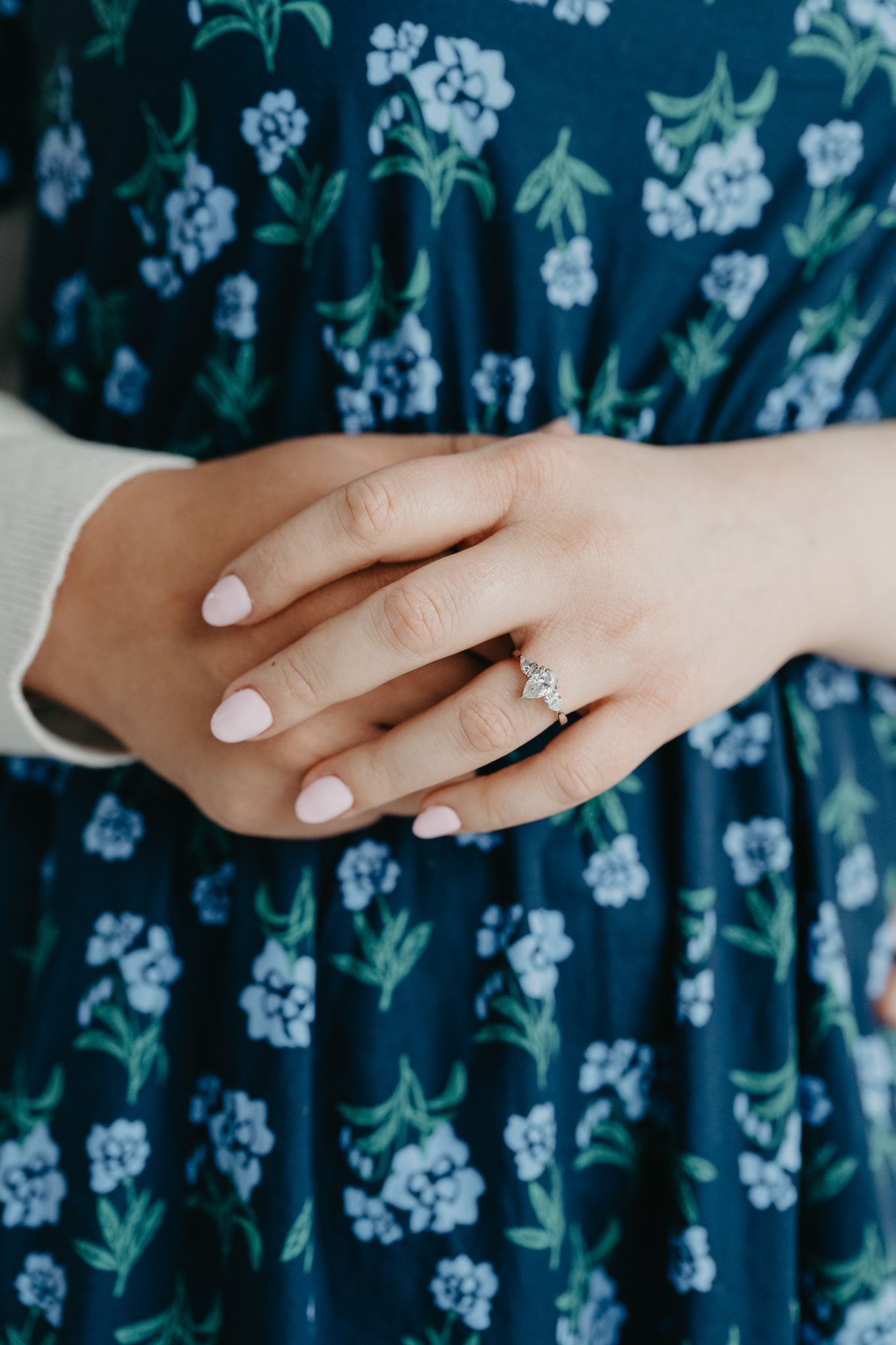 A person wearing a blue floral dress with their hands clasped, highlighting a diamond ring on their ring finger.