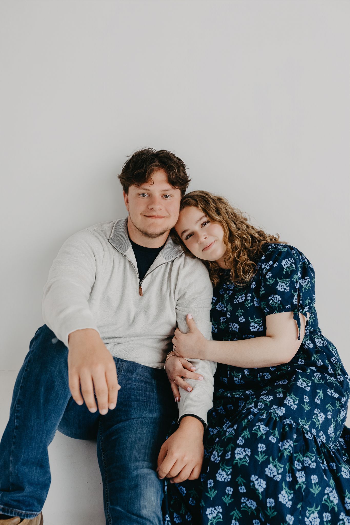 A young couple poses against a white wall; the man wears a light sweater and jeans, the woman a blue floral dress.
