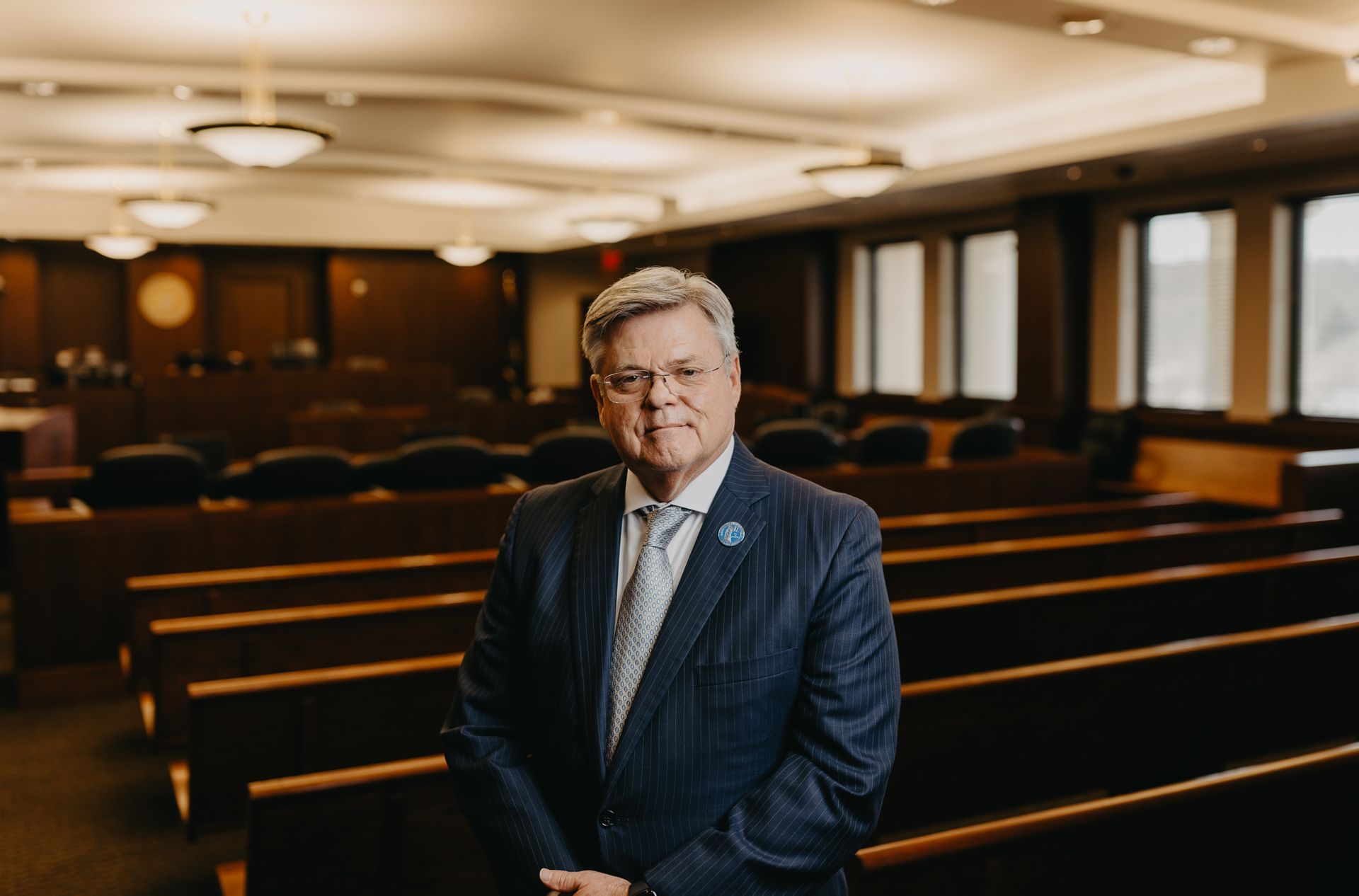 A man in a suit and tie stands in a courtroom, facing the camera with a neutral expression.