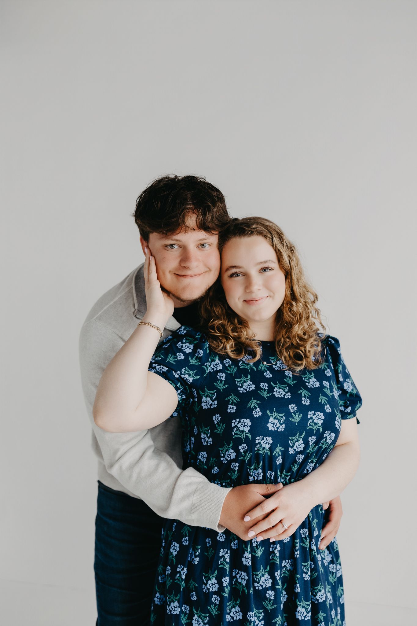 A person wearing a grey sweater embraces a person in a blue floral dress from behind against a plain white background.