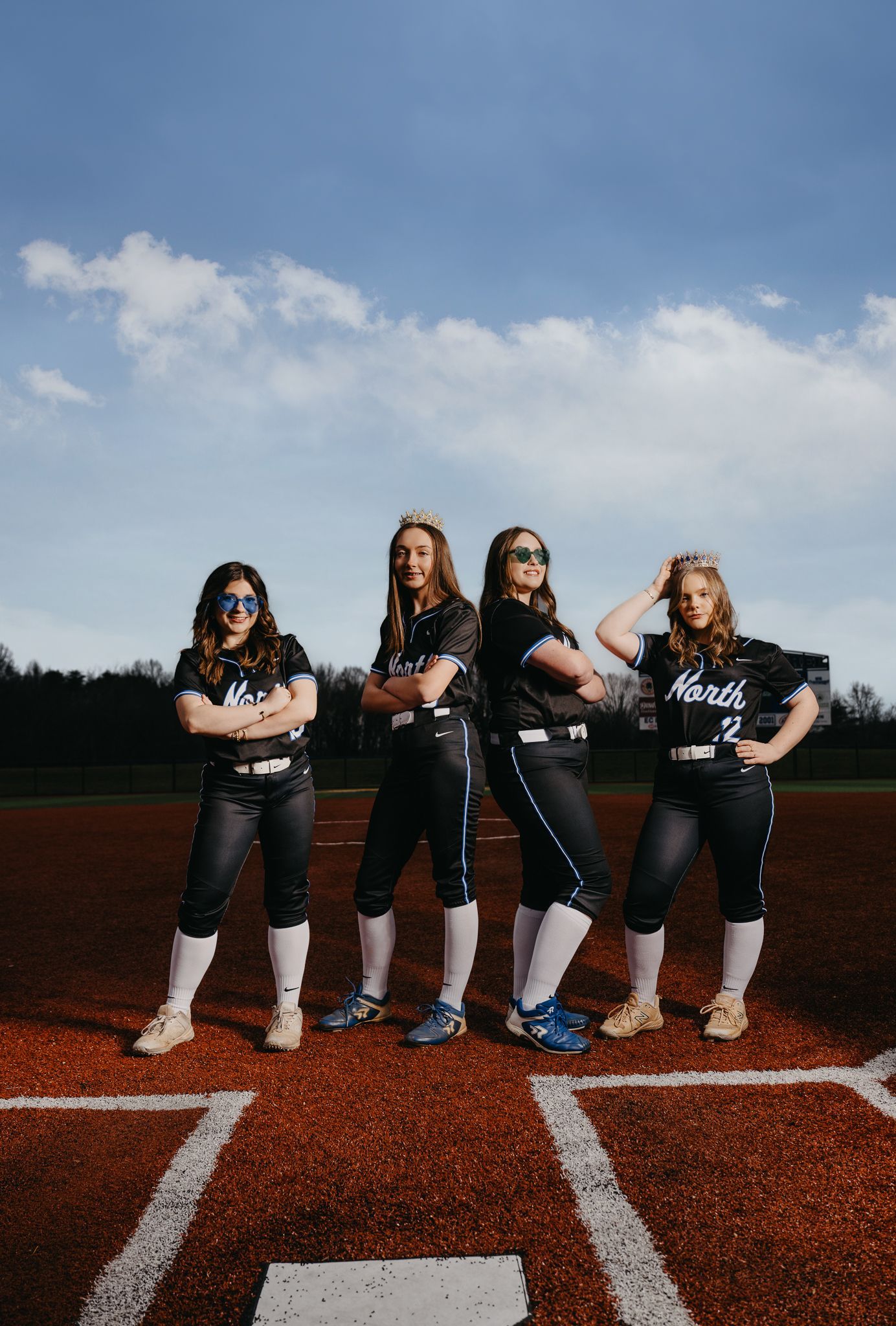 Four softball players in black uniforms stand in a row on a dirt field with white base lines under a cloudy blue sky.