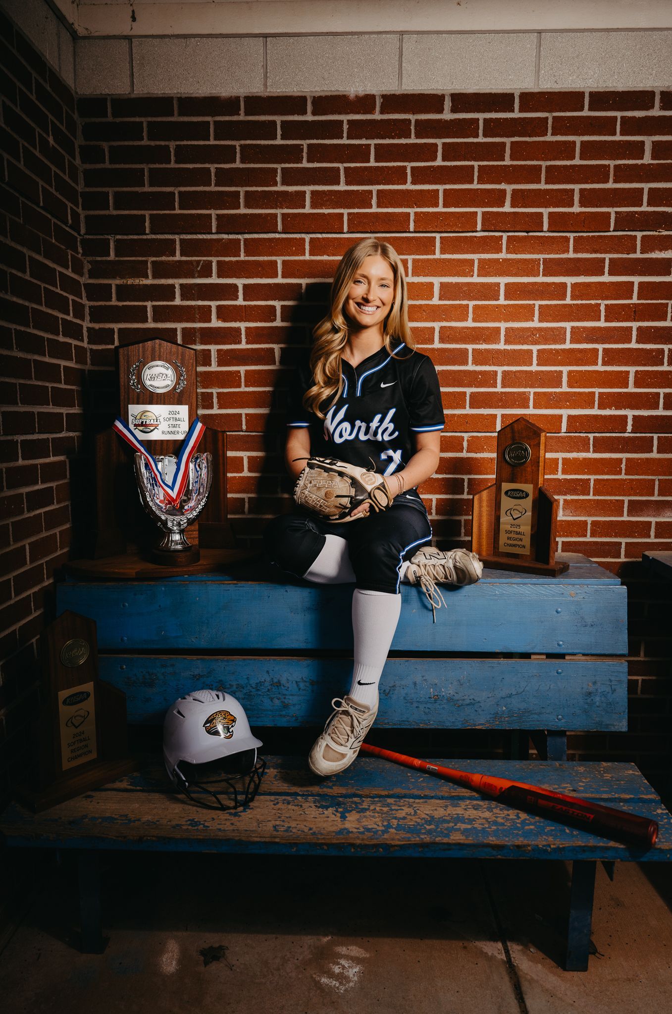 A softball player sits on a blue dugout bench in front of a brick wall, surrounded by trophies, a helmet, and a bat.