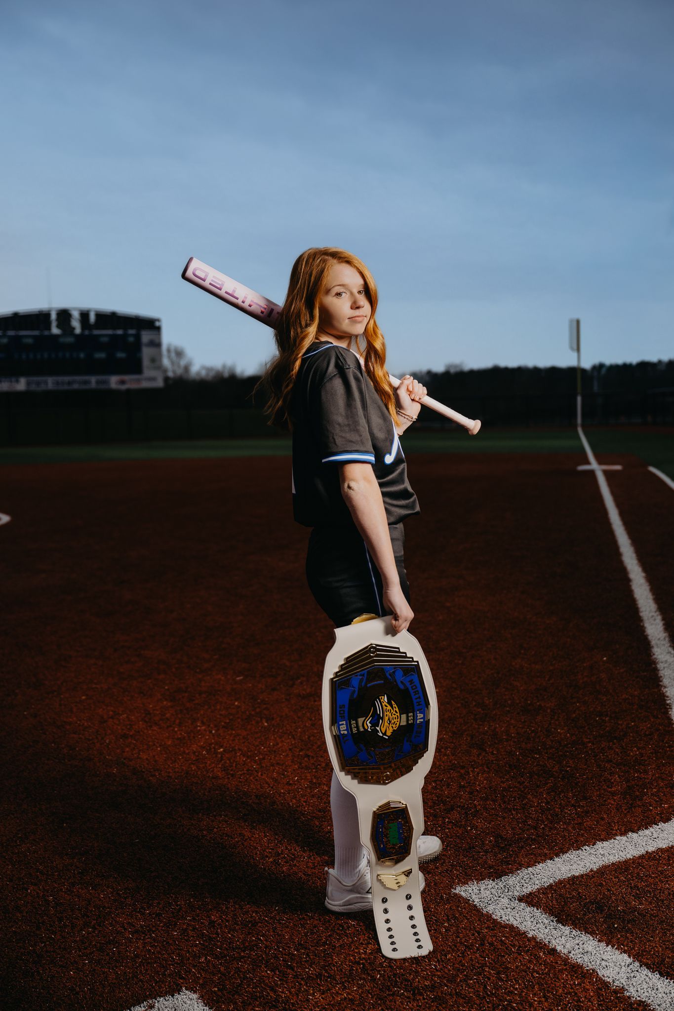 A person on a baseball field holds a bat on their shoulder and a championship belt in their hand, looking at the camera.