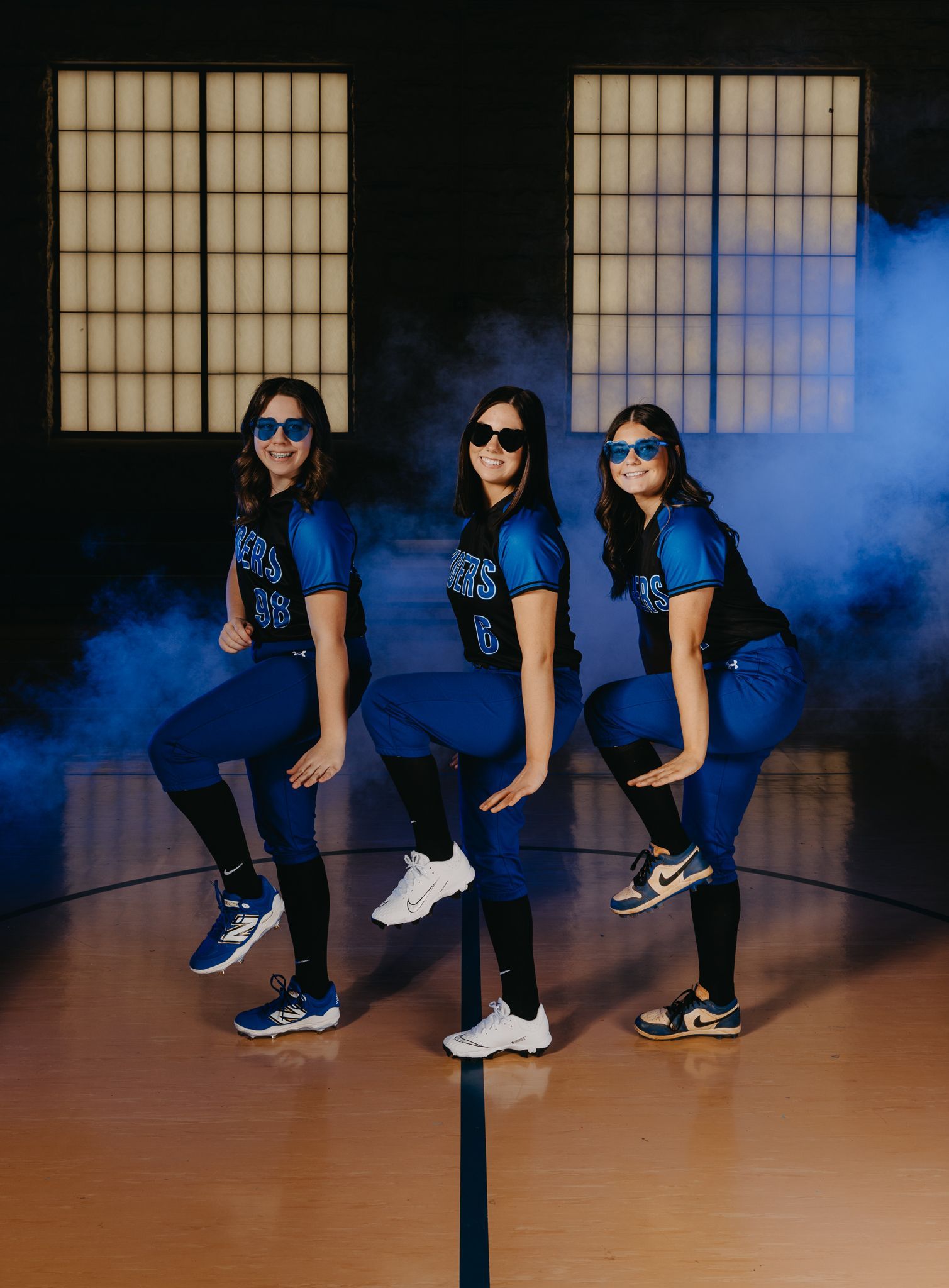 Three people wearing blue athletic uniforms and sunglasses strike a matching pose on a basketball court in blue fog.