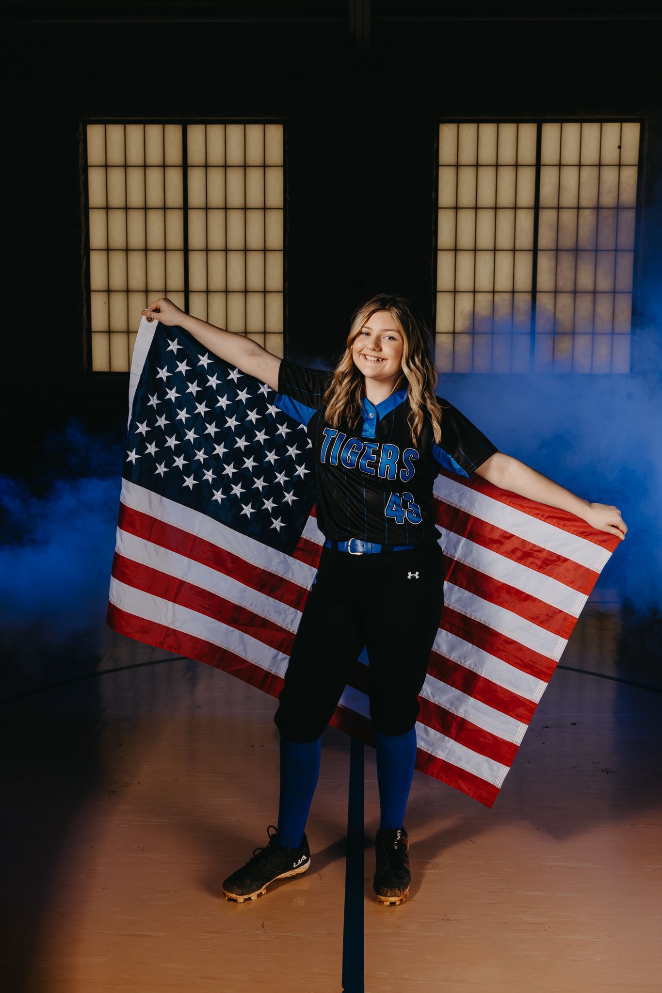 A smiling person in a blue and black softball uniform stands indoors holding a large American flag spread behind them.