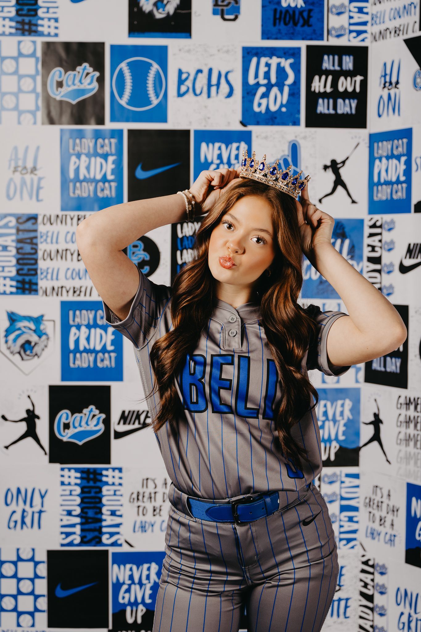 A person in a gray baseball uniform wearing a crown, posing in front of a blue and white patterned wall.