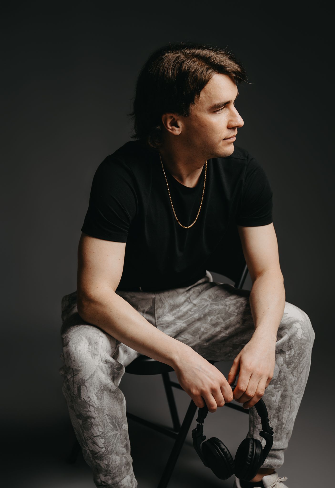 A person sits on a chair, wearing a black t-shirt and grey patterned joggers, holding headphones against a grey background.