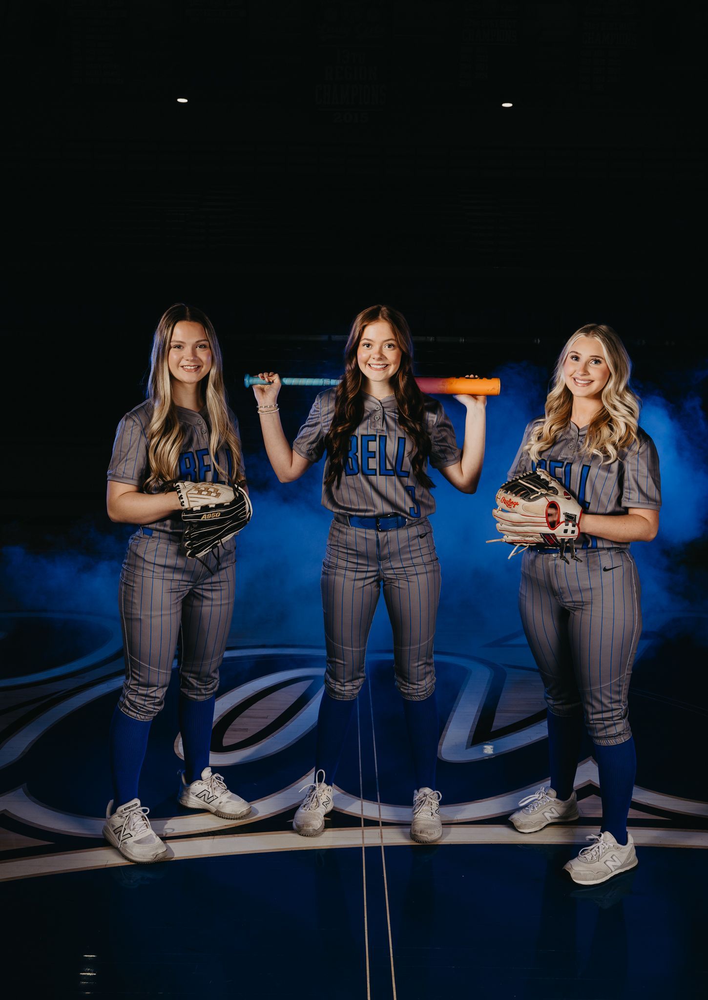 Three softball players in gray uniforms pose together with gloves and a bat against a blue, smoky, indoor background.