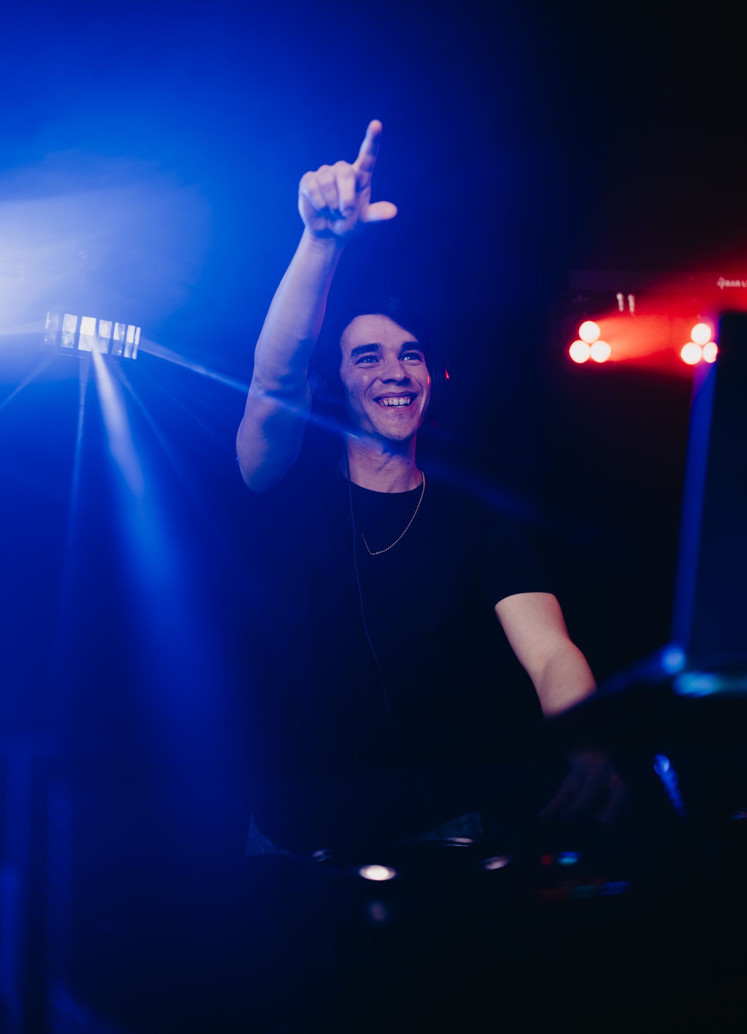 A person smiles while pointing upward from behind a DJ console, illuminated by bright blue stage lights.