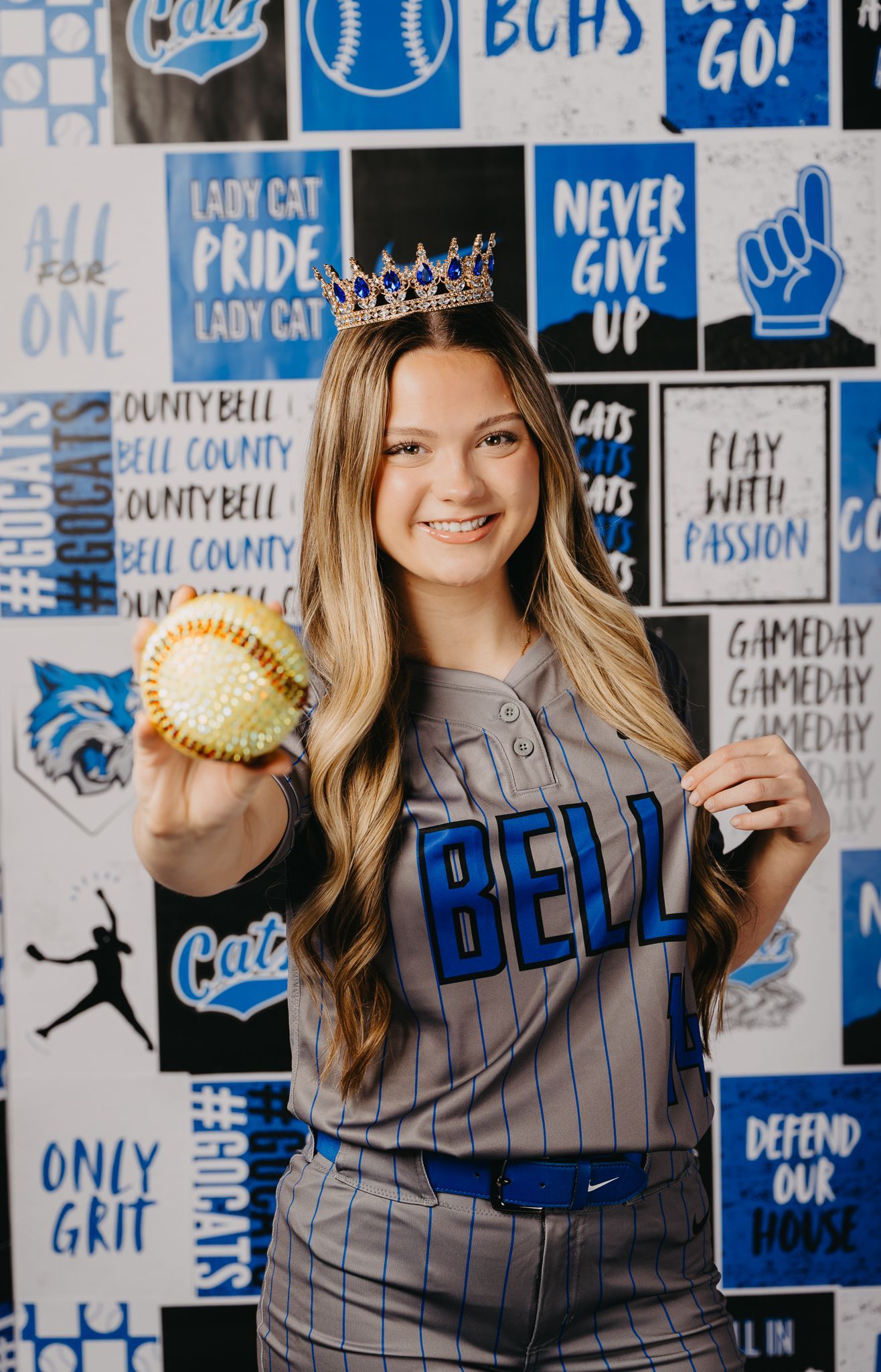 A smiling softball player wearing a crown and grey uniform holds a yellow ball in front of a blue and white sports banner.