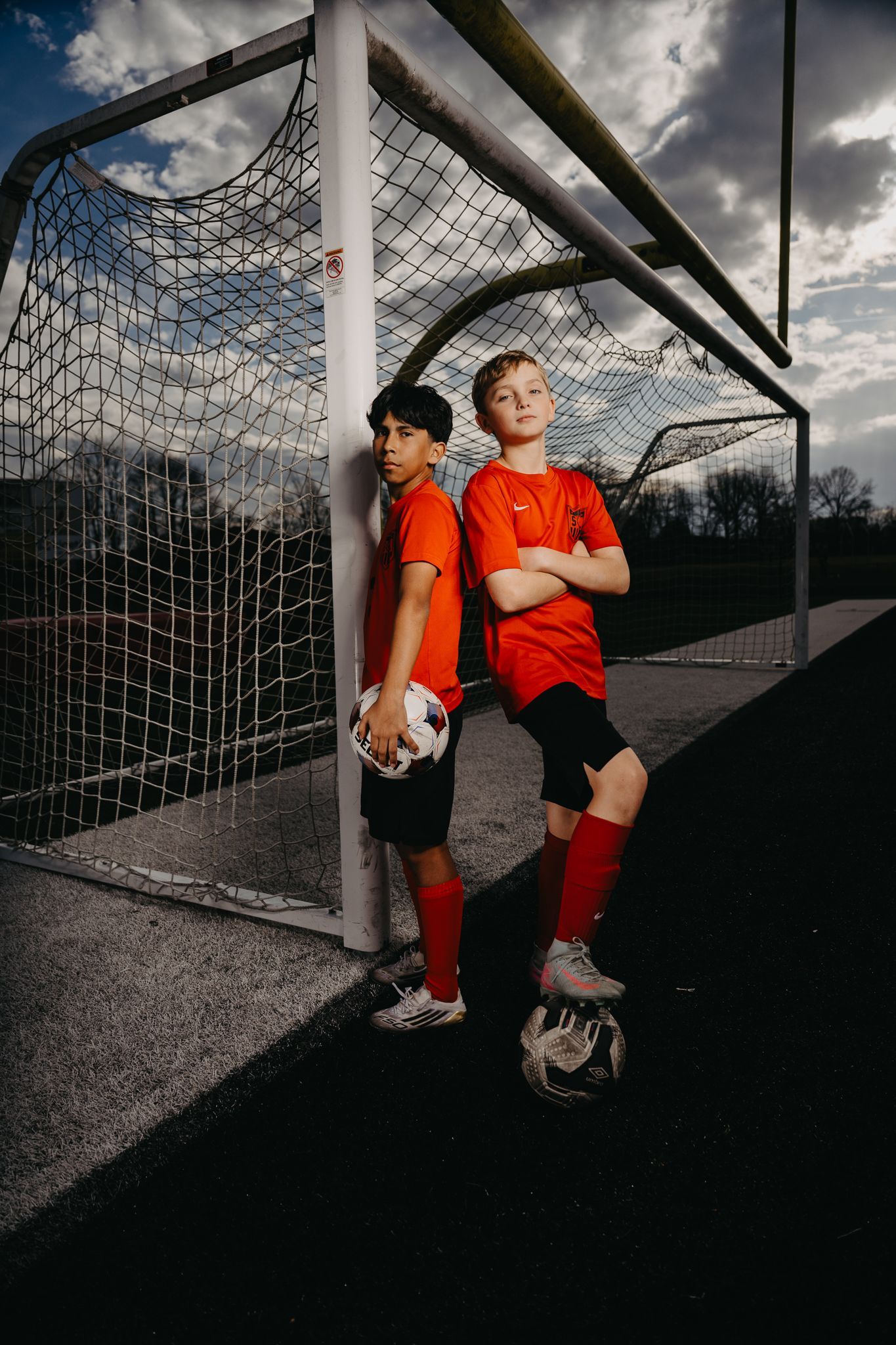 Two players in orange uniforms stand back-to-back by a soccer goal on a field, one holding a soccer ball.