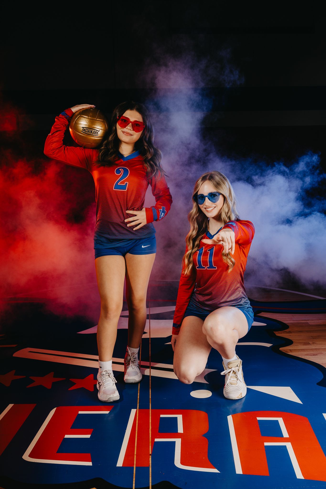 Two athletes in red and blue uniforms with sunglasses pose with a sports ball amid red and blue smoke on a gym floor.