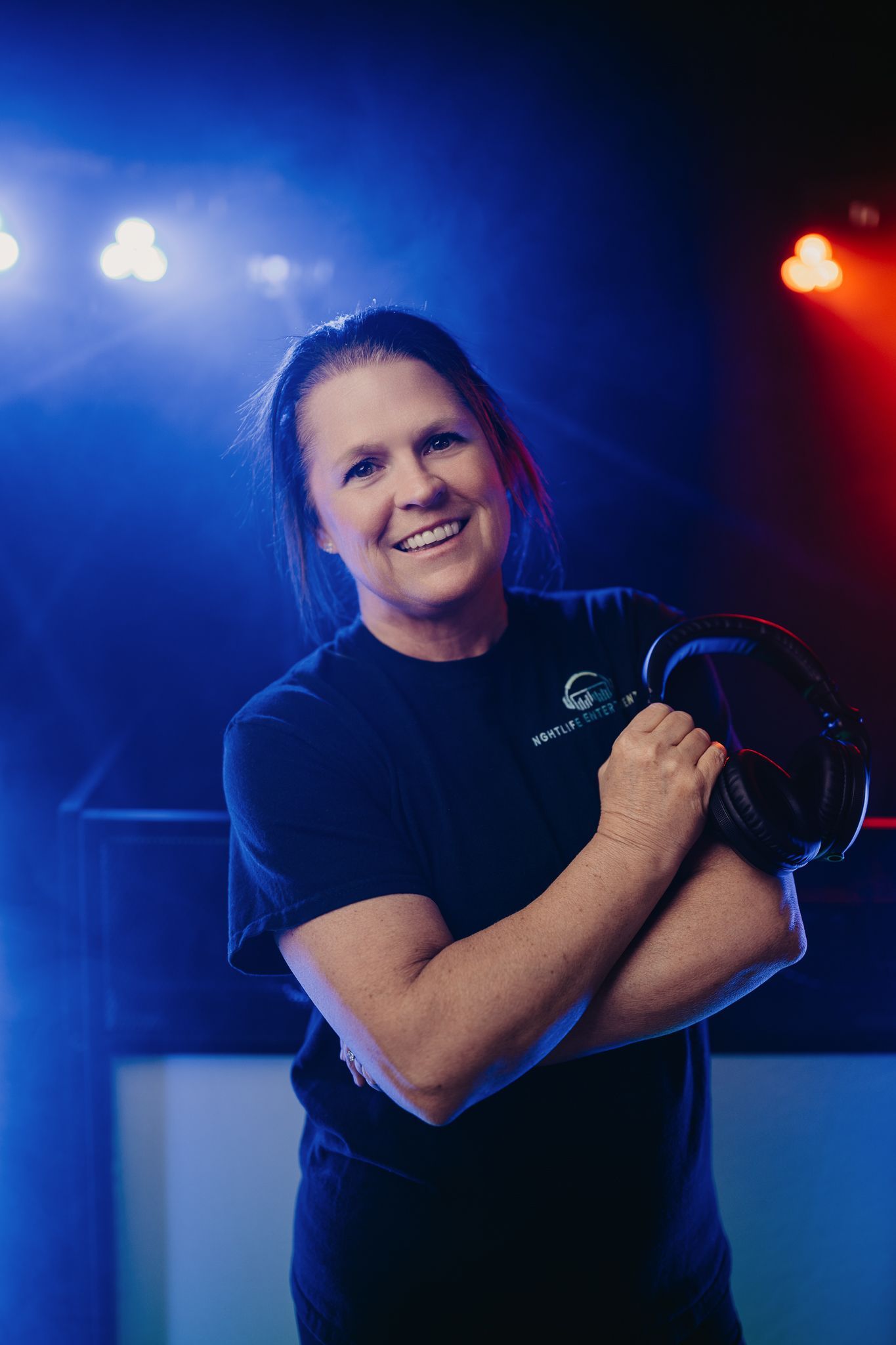 A smiling person wearing a black shirt and holding headphones stands in front of a stage with blue and red lighting.