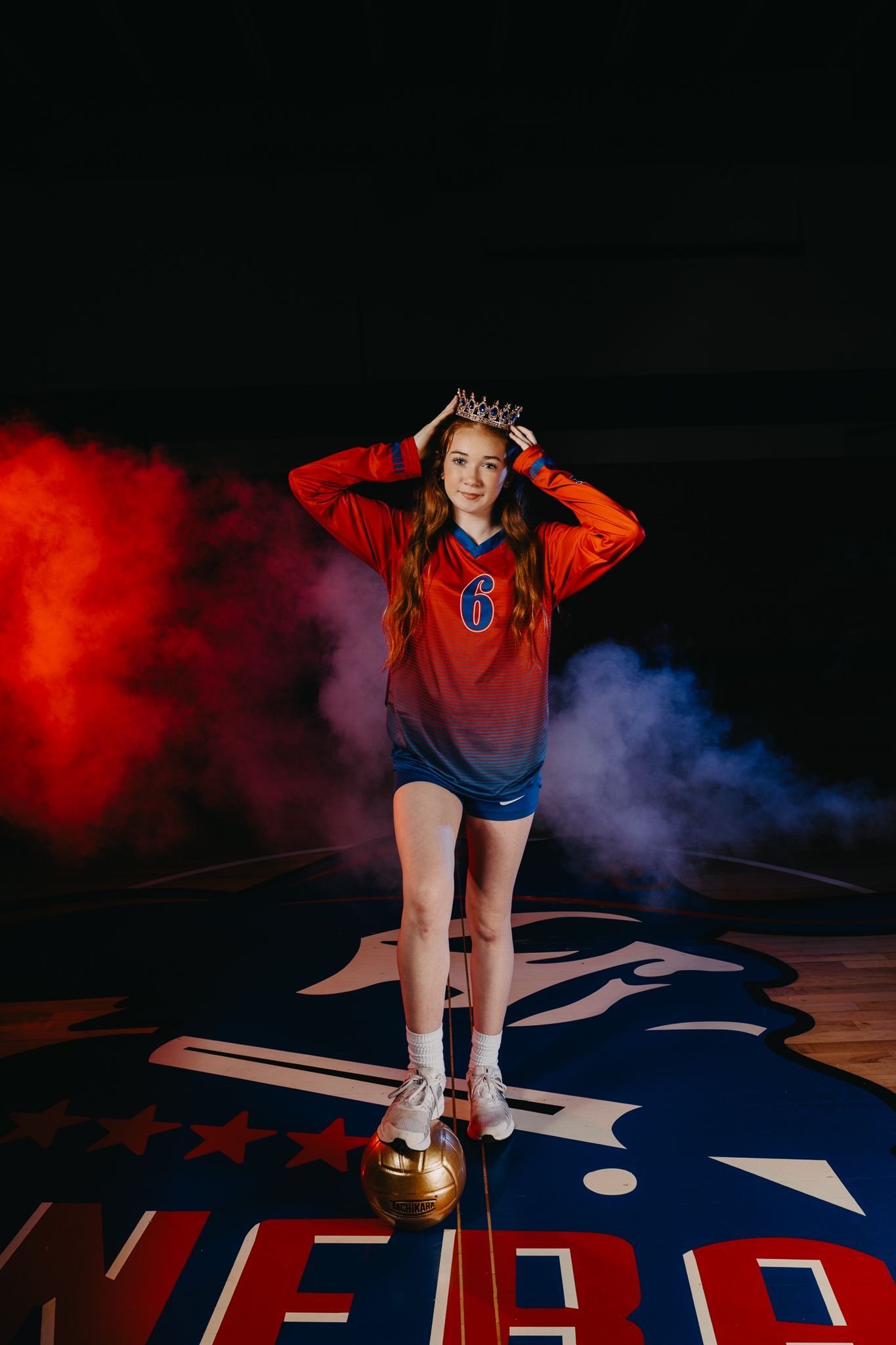 A player in an orange and blue jersey stands on a basketball court with a ball, framed by red and blue smoke.