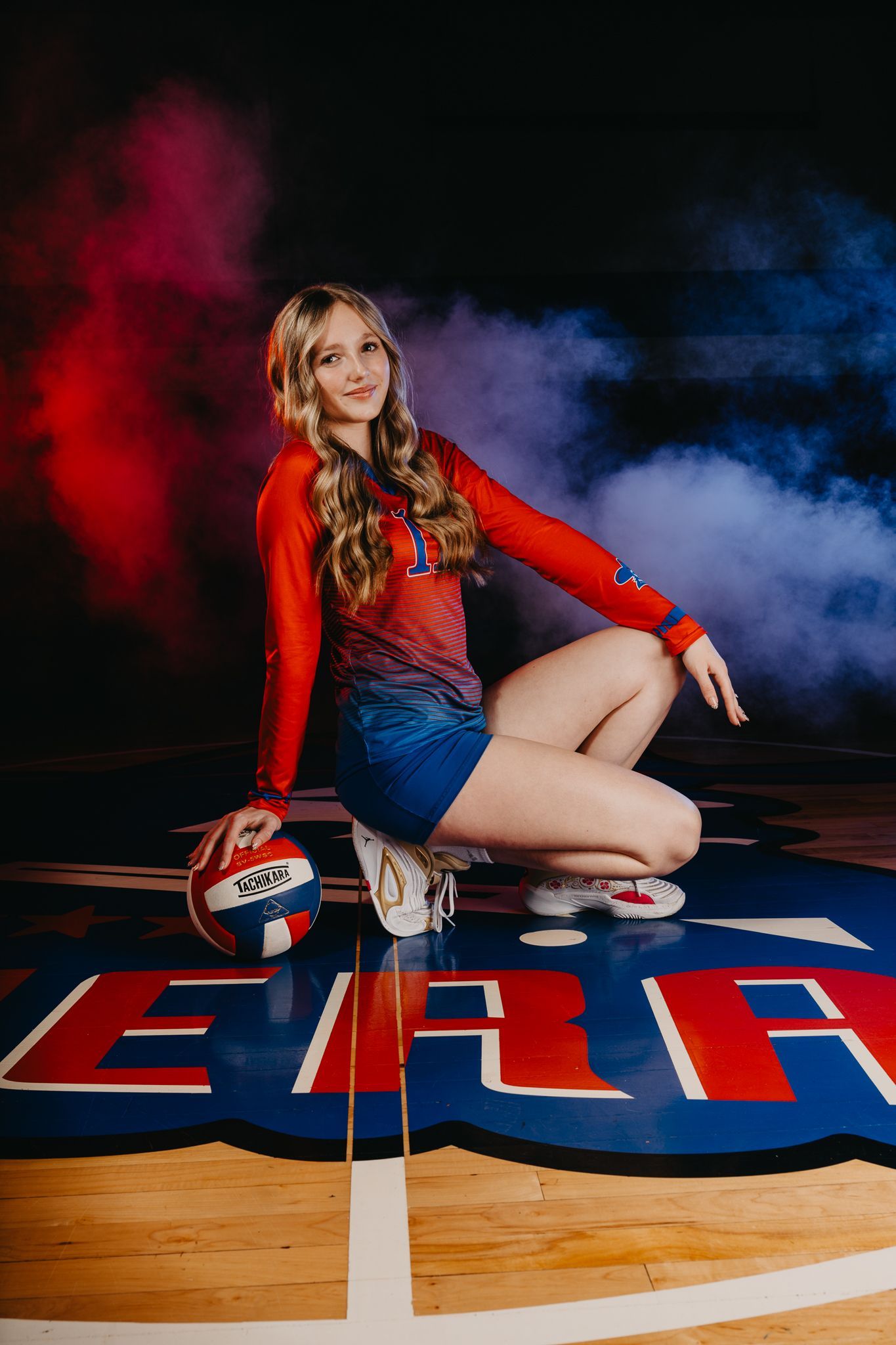 A person in a red and blue volleyball uniform crouching on a court, resting a hand on a volleyball with smoke in background.