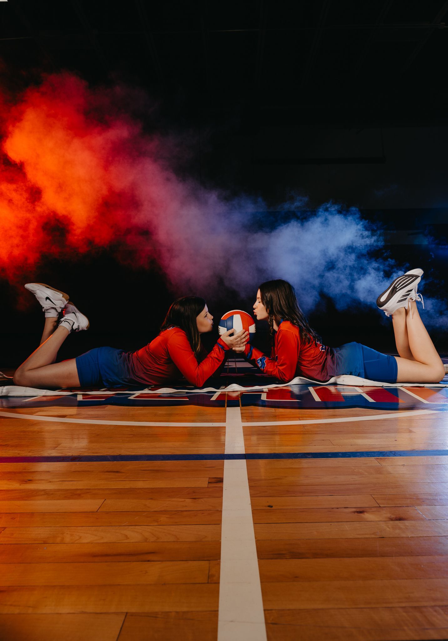 Two volleyball players lie on a court, face-to-face, holding a ball between them amid swirling red and blue smoke.