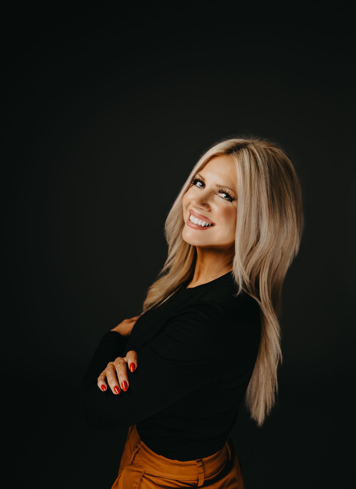 A smiling person with long, textured blonde hair and a black long-sleeved shirt posing with arms crossed against black.