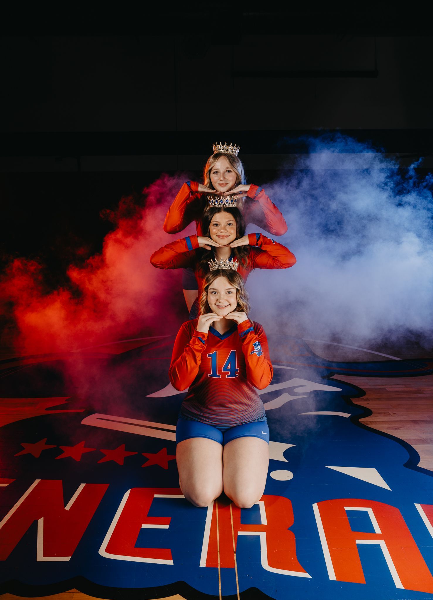 Three cheerleaders in red and blue uniforms are stacked vertically, kneeling on a gym floor amidst red and blue smoke.