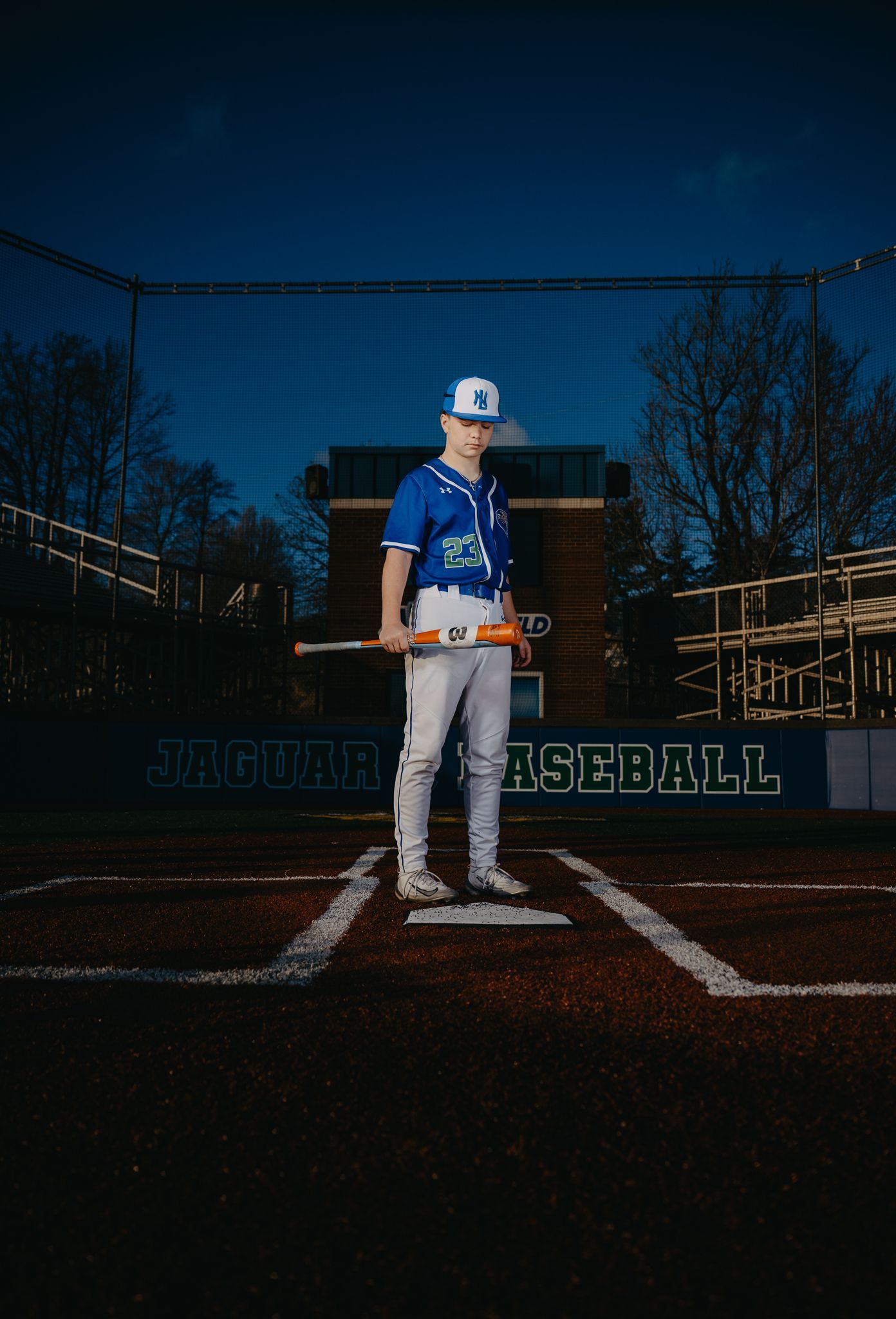 A baseball player in a blue uniform stands on a dirt field holding a bat, with a baseball backstop visible behind them.