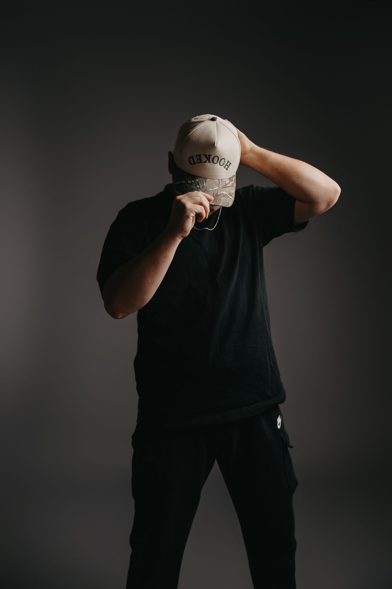 A person in a black t-shirt and cargo pants standing against a dark grey background, pulling down a light-colored cap.